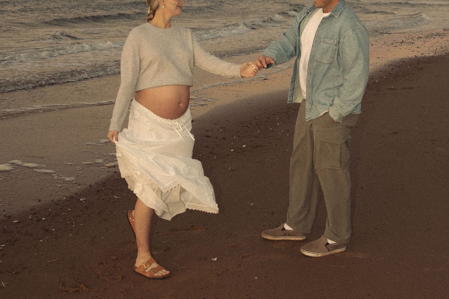 A couple playfully interacts on a dark sandy beach at sunset.