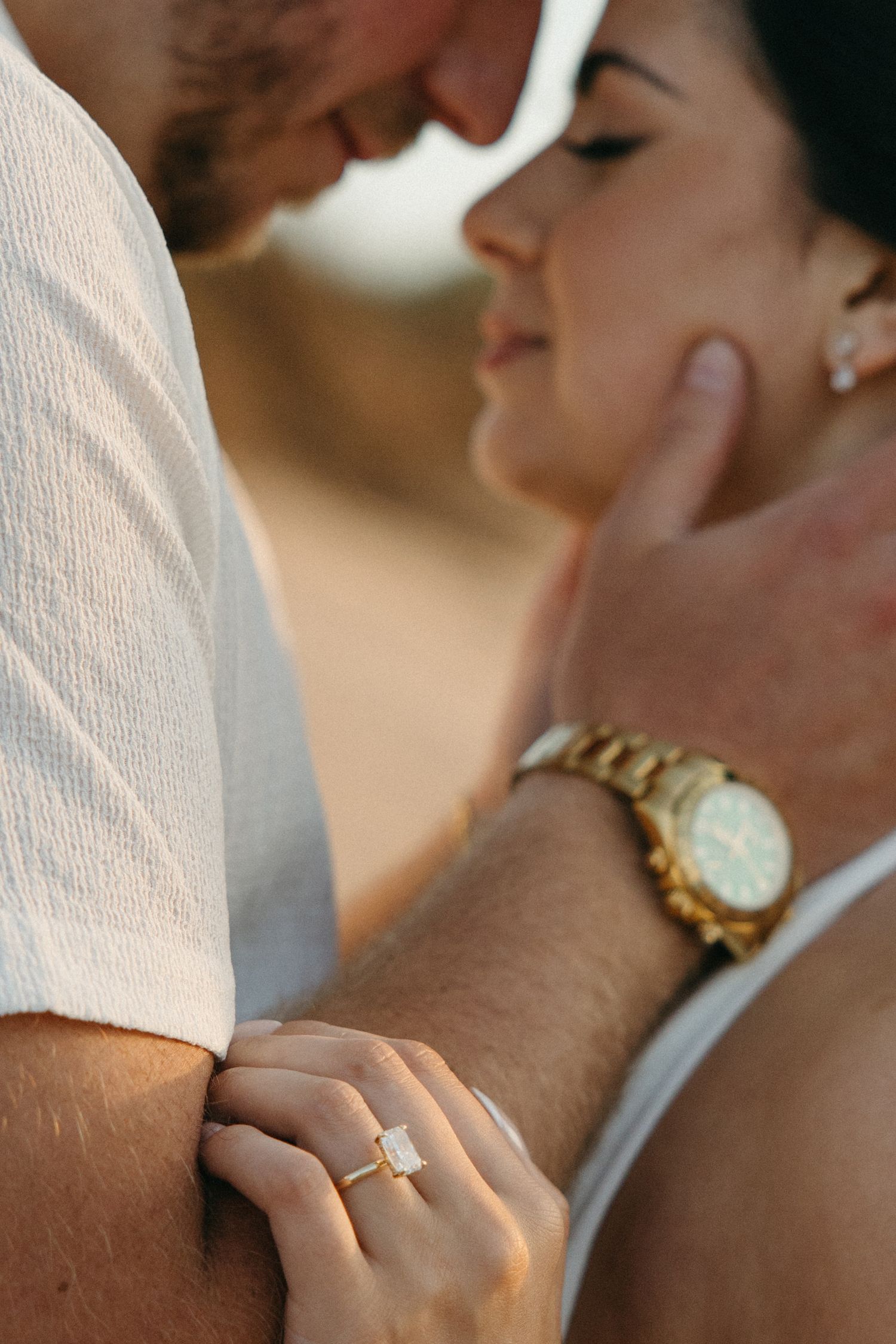 Intimate close-up of couple almost kissing, woman displaying emerald-cut engagement ring and gold watch.