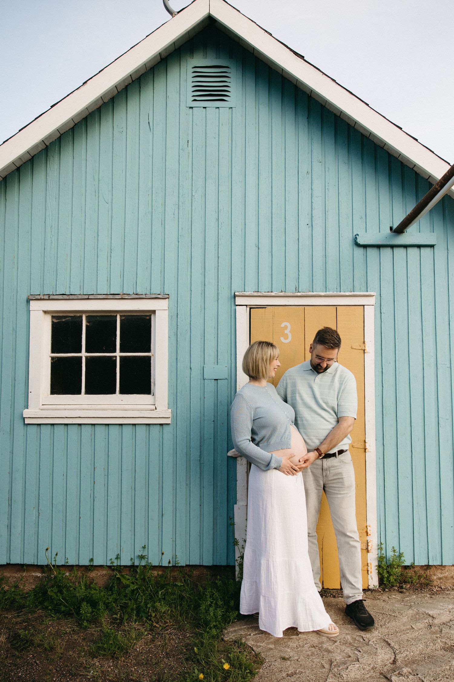 A couple holds hands in front of a light blue wooden building with a yellow door.