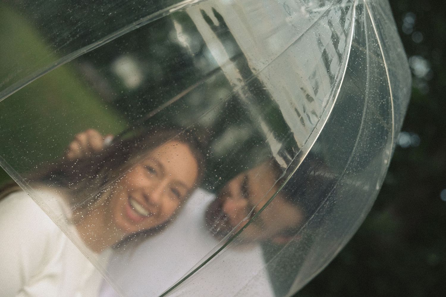 Couple smiling together under a clear transparent umbrella in the rain, captured in a romantic outdoor portrait.