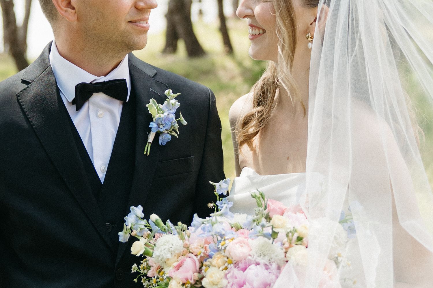 Bride and groom in black tuxedo with blue boutonniere smiling at each other holding pastel wedding bouquet.