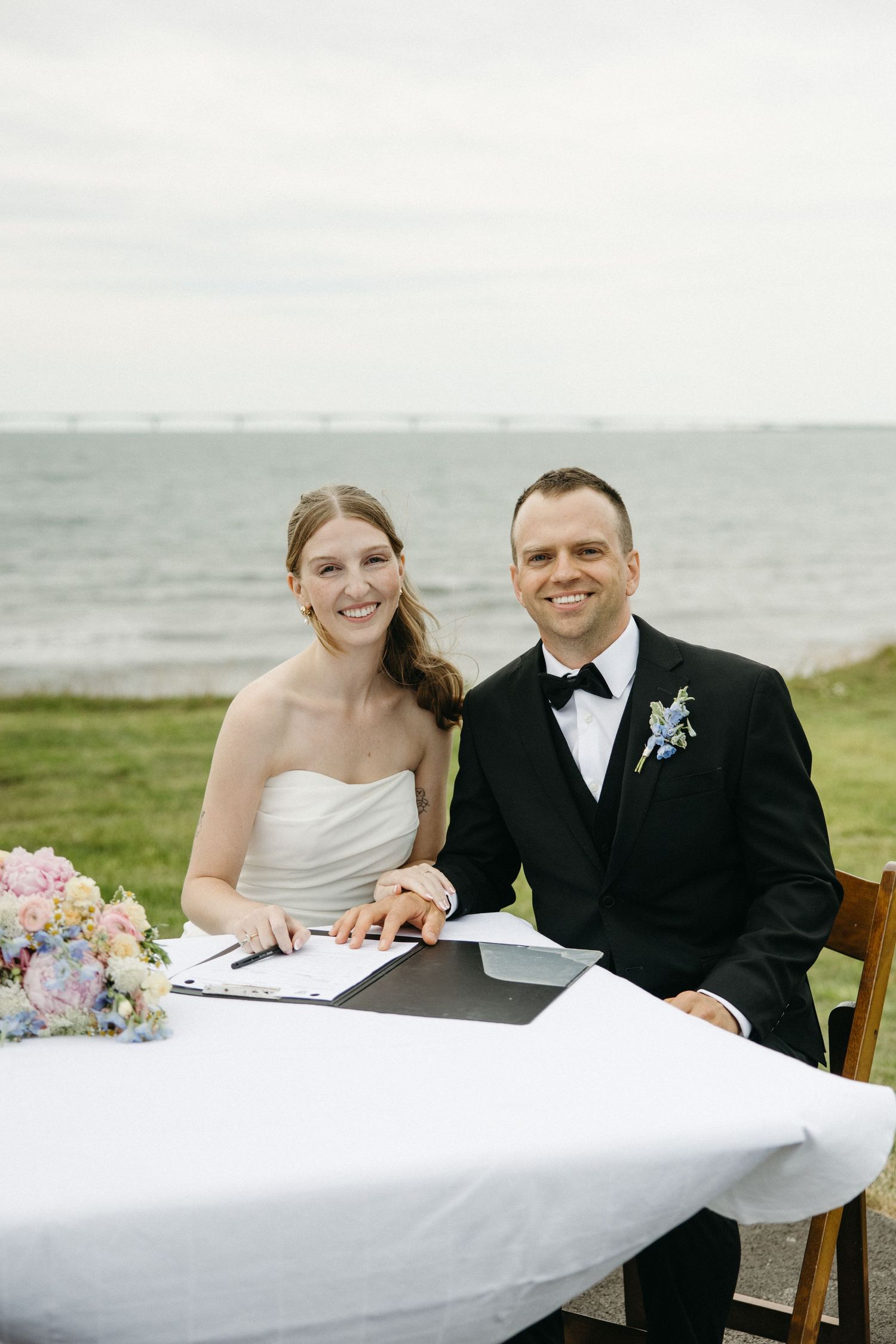 Smiling bride and groom signing marriage documents at a waterfront table with floral bouquet.