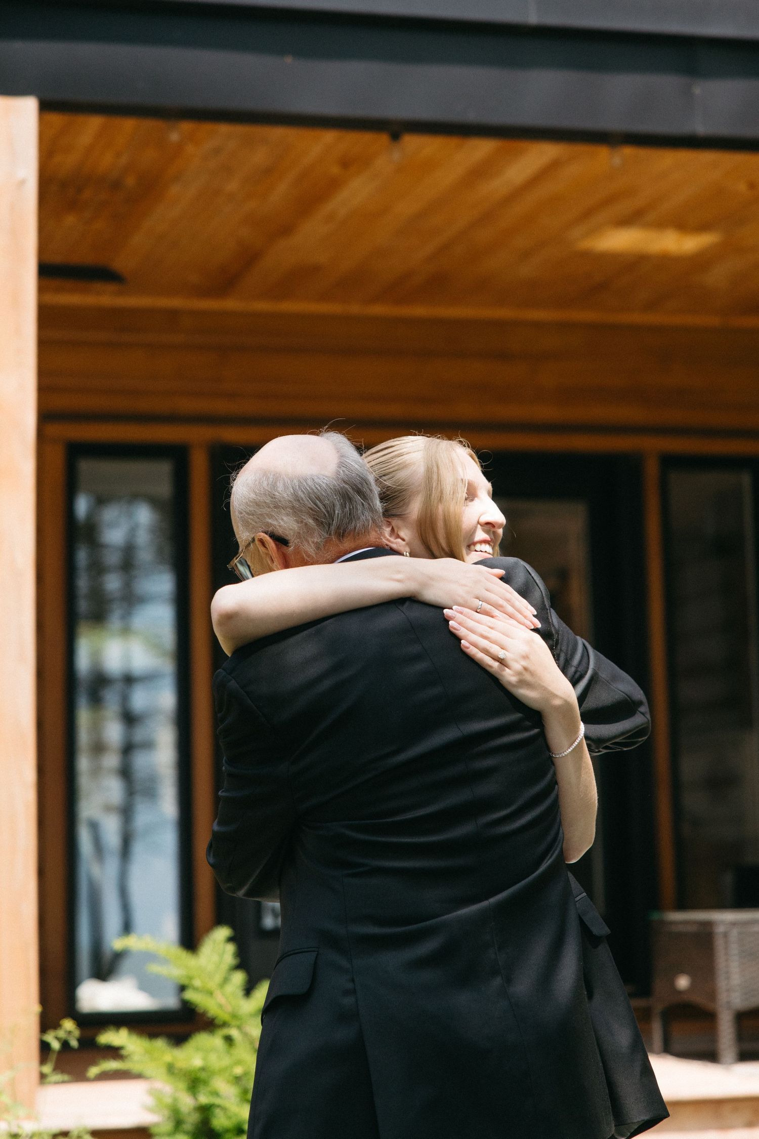 Bride and older gentleman sharing warm embrace outside modern wooden venue building on wedding day.