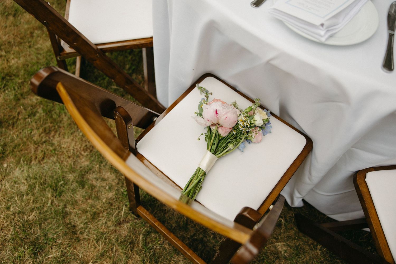 Delicate pink and white bridal bouquet rests on a white cushion of a wooden folding chair at wedding.