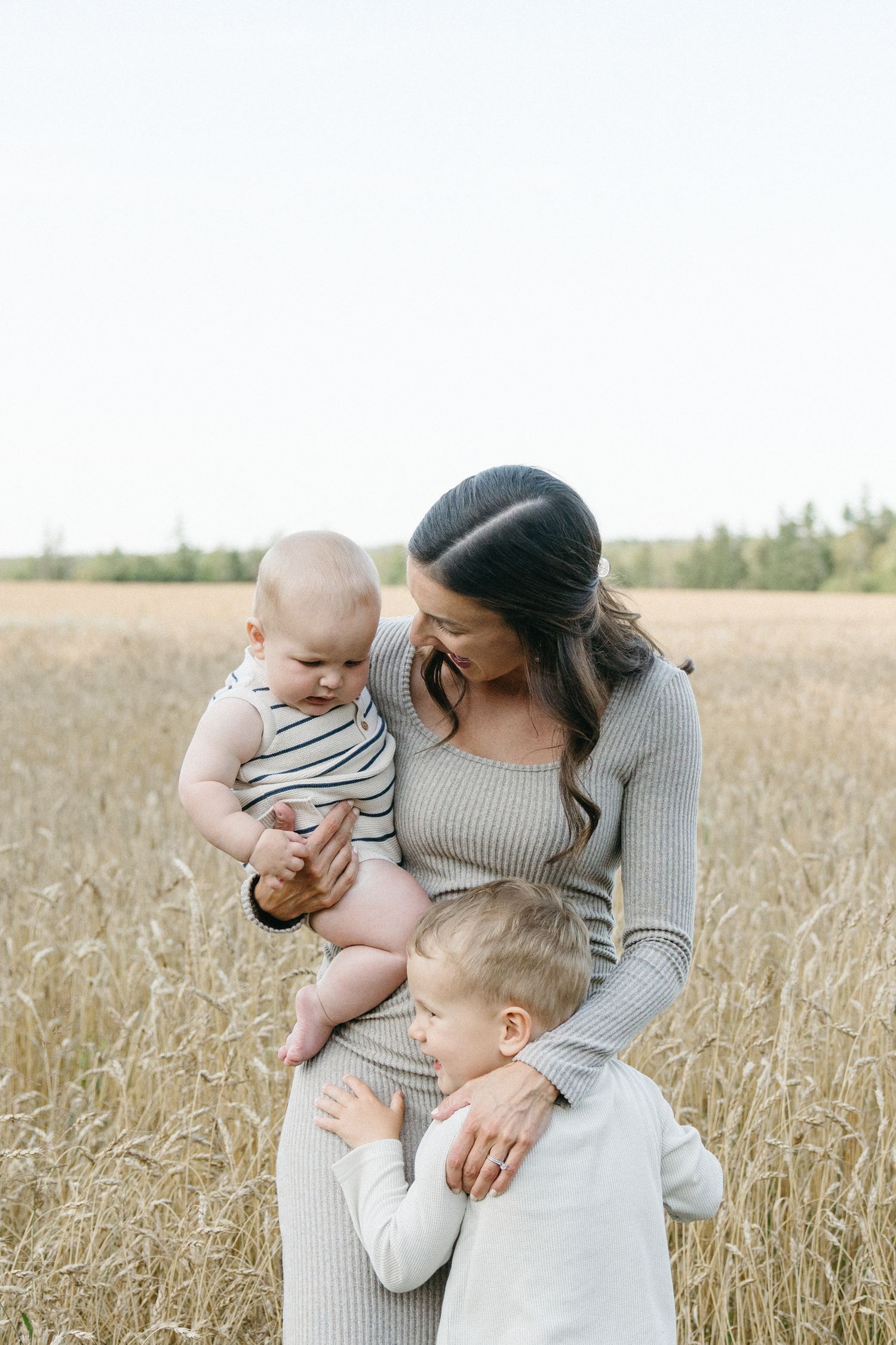 A mother holds her baby while her toddler hugs her in a golden wheat field.
