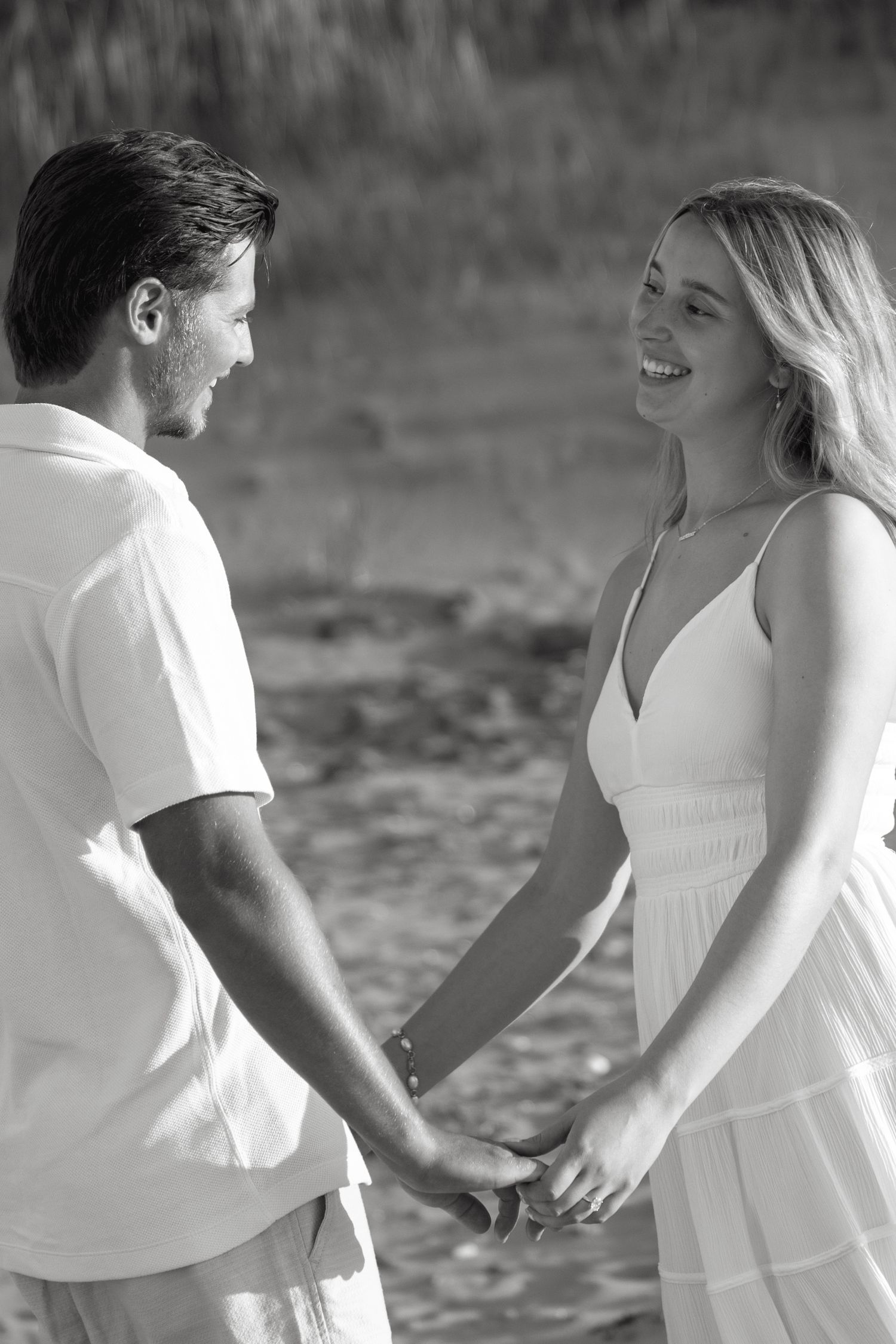Black and white photo of a smiling couple holding hands outdoors, woman in white dress, man in white shirt.