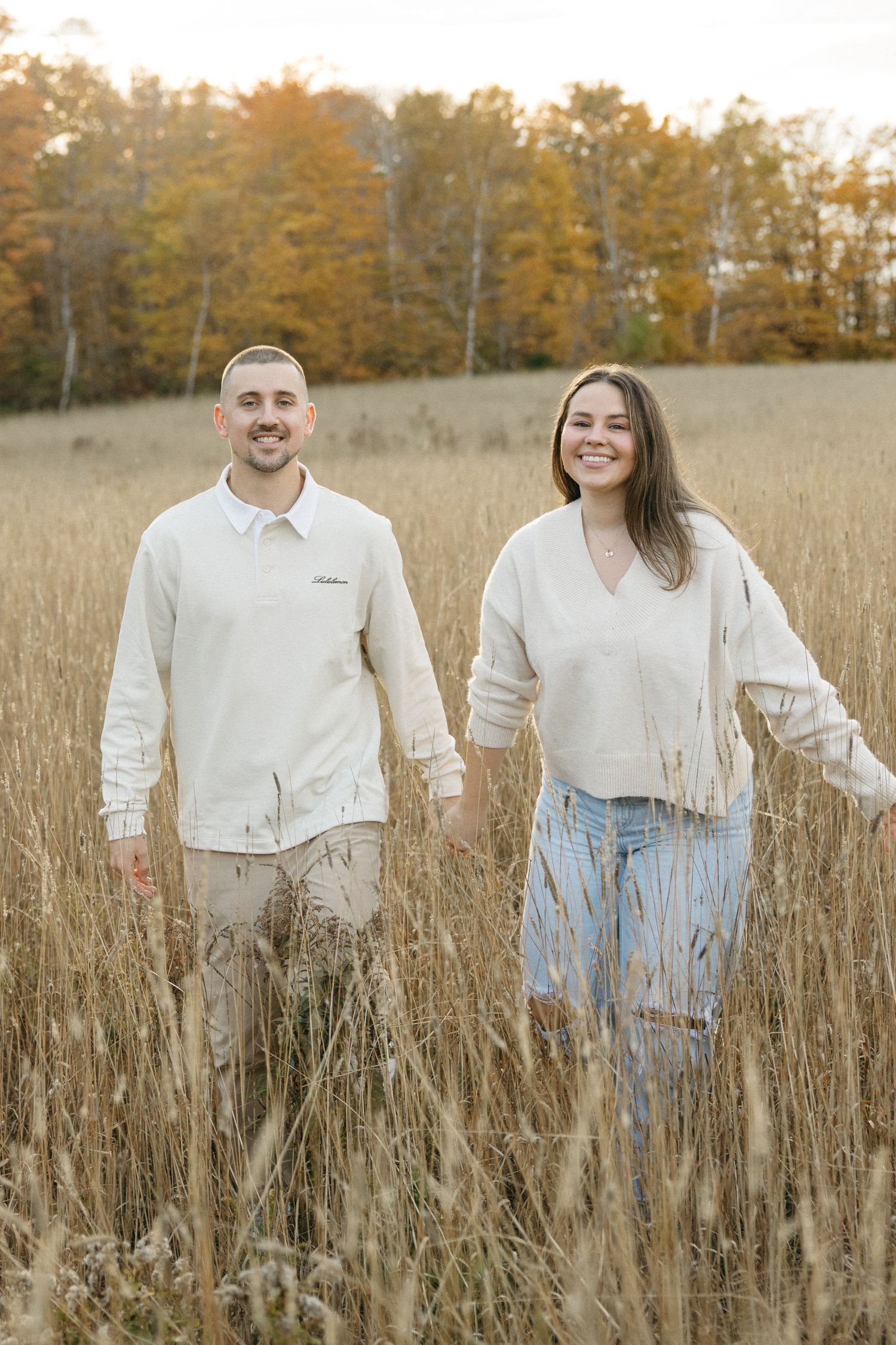 Two people in matching white tops and jeans stand among tall autumn grasses.