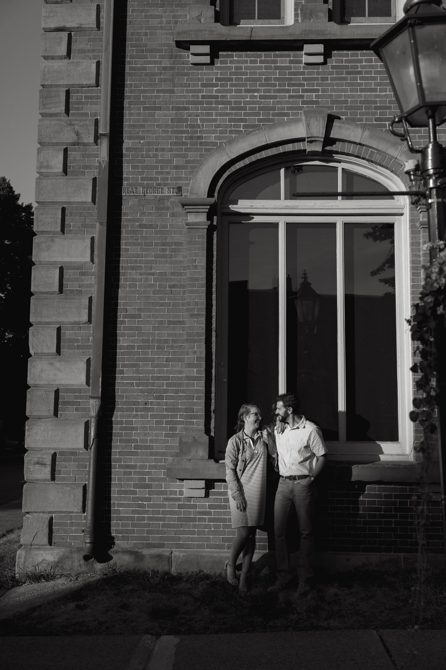 A couple stands together in front of a historic brick building with arched doorway.