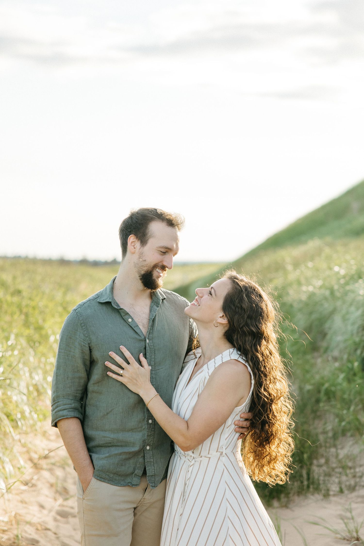 A smiling couple embraces on a sandy path surrounded by green dunes, the woman in a striped dress gazing up at the man.