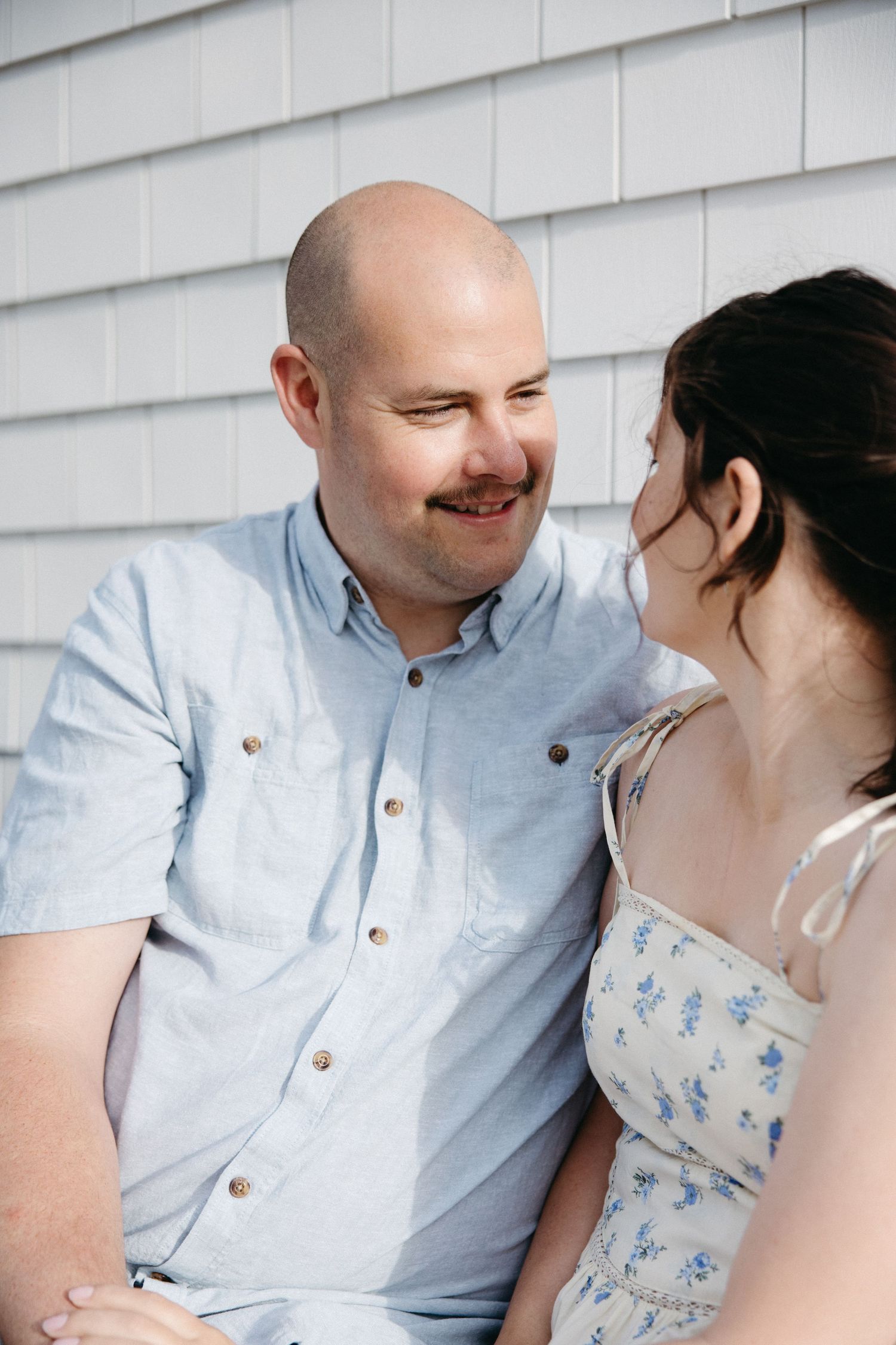 A bald man with a mustache in a light blue shirt smiles at a dark-haired woman in a floral dress outdoors.
