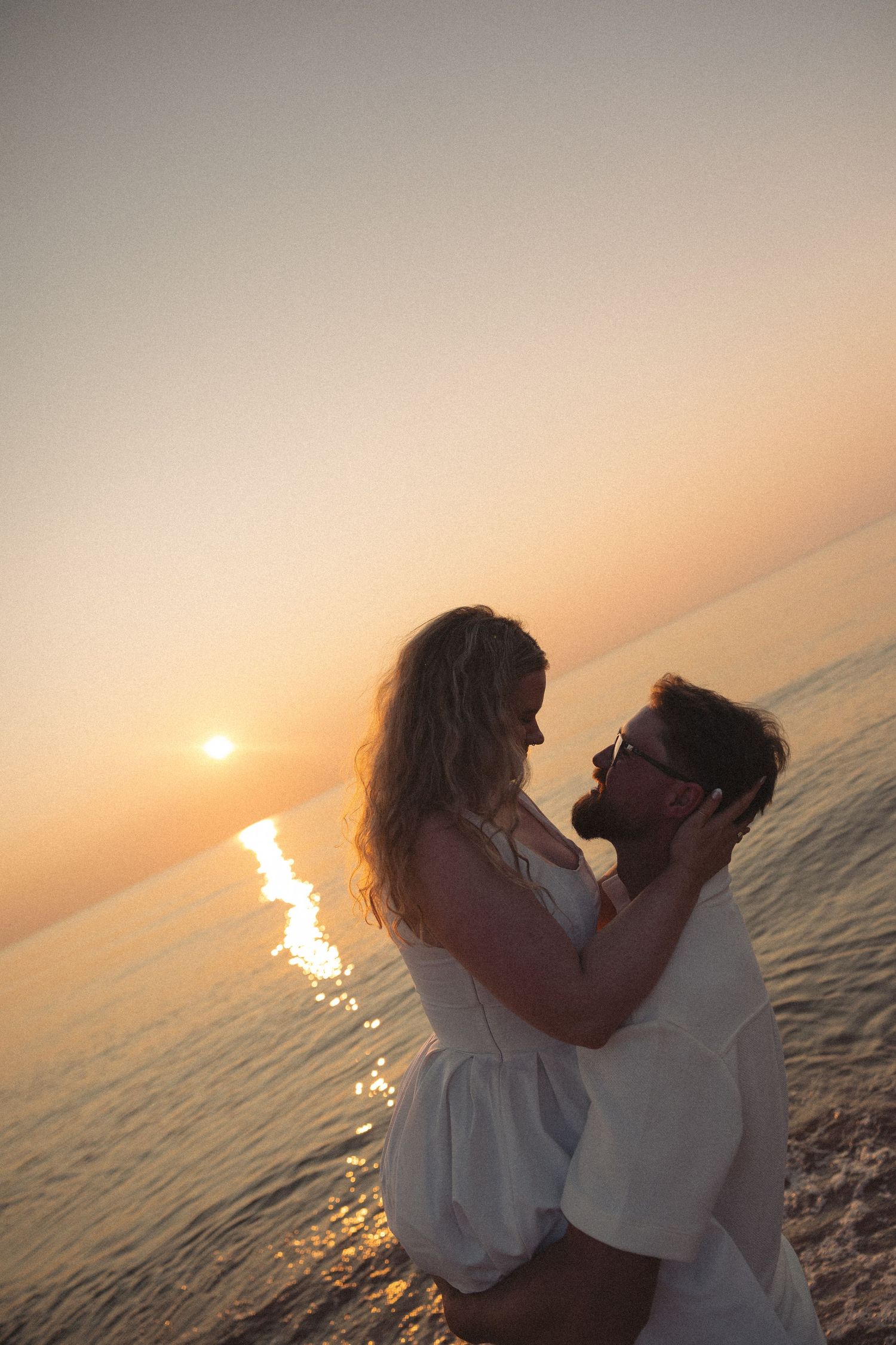 Romantic couple embracing at golden sunset over calm ocean waters, both dressed in white.