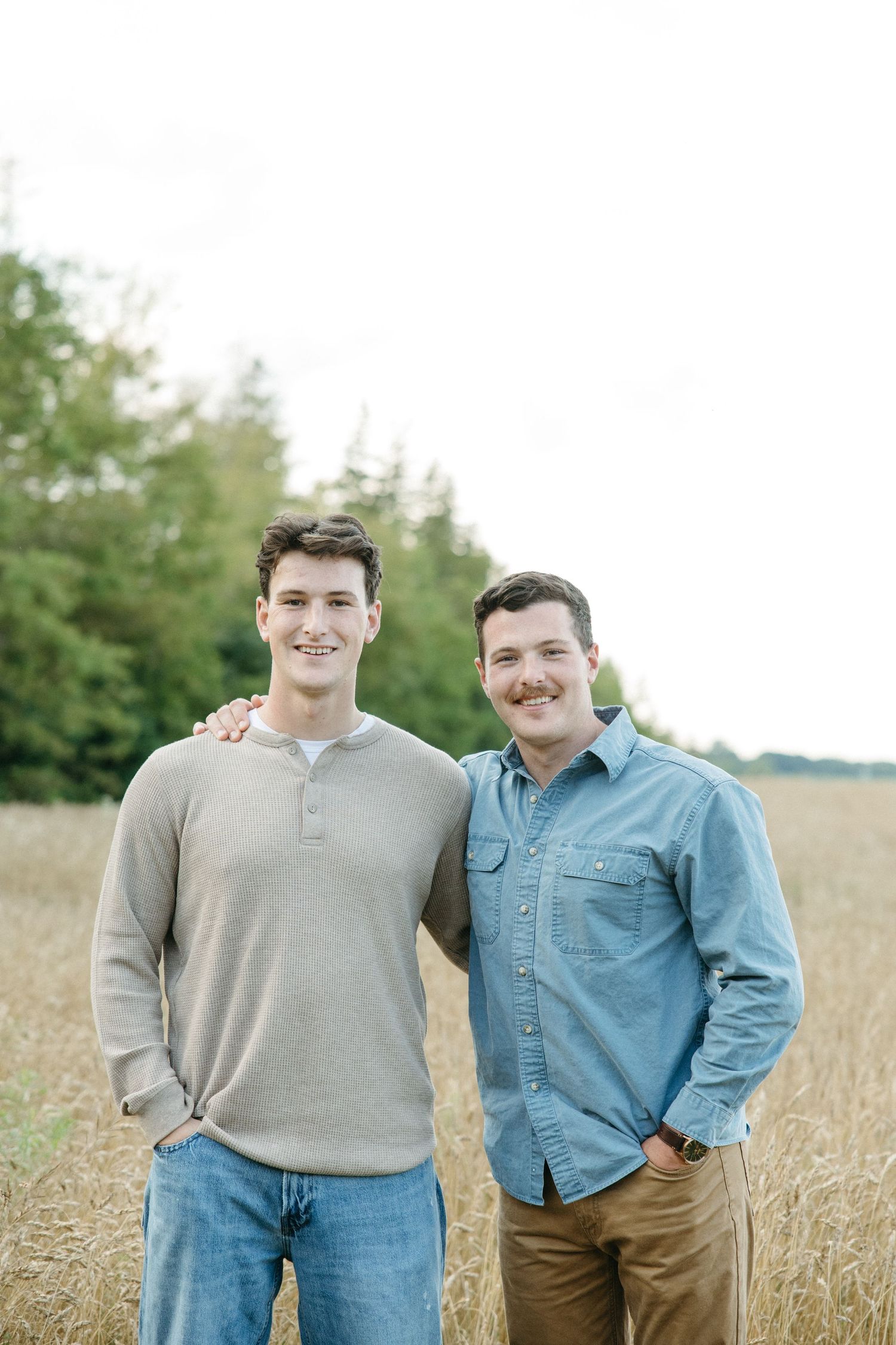 Two young men smiling outdoors in a golden wheat field with trees in the background.