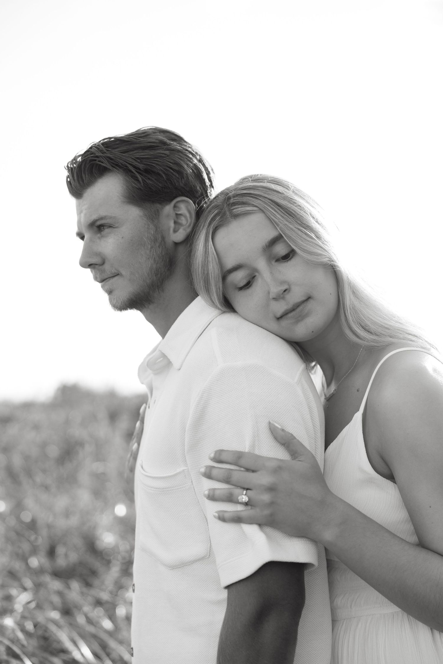 Black and white portrait of woman embracing man from behind in field at sunset.