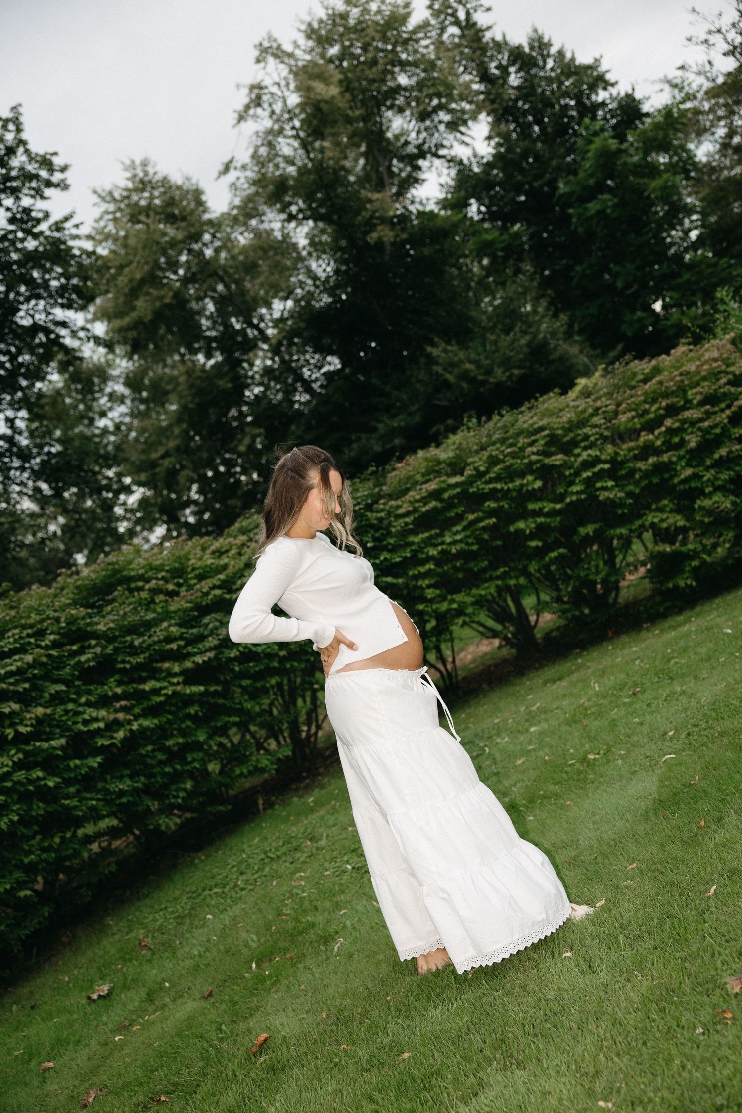 Pregnant woman in white long-sleeve maxi dress stands in lush green park, cradling baby bump.