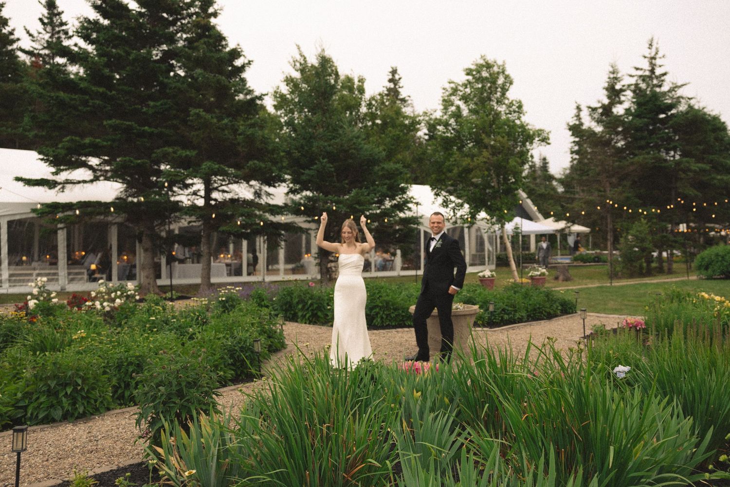 Bride and groom walking through a lush garden venue with a white tent and string lights in the background.
