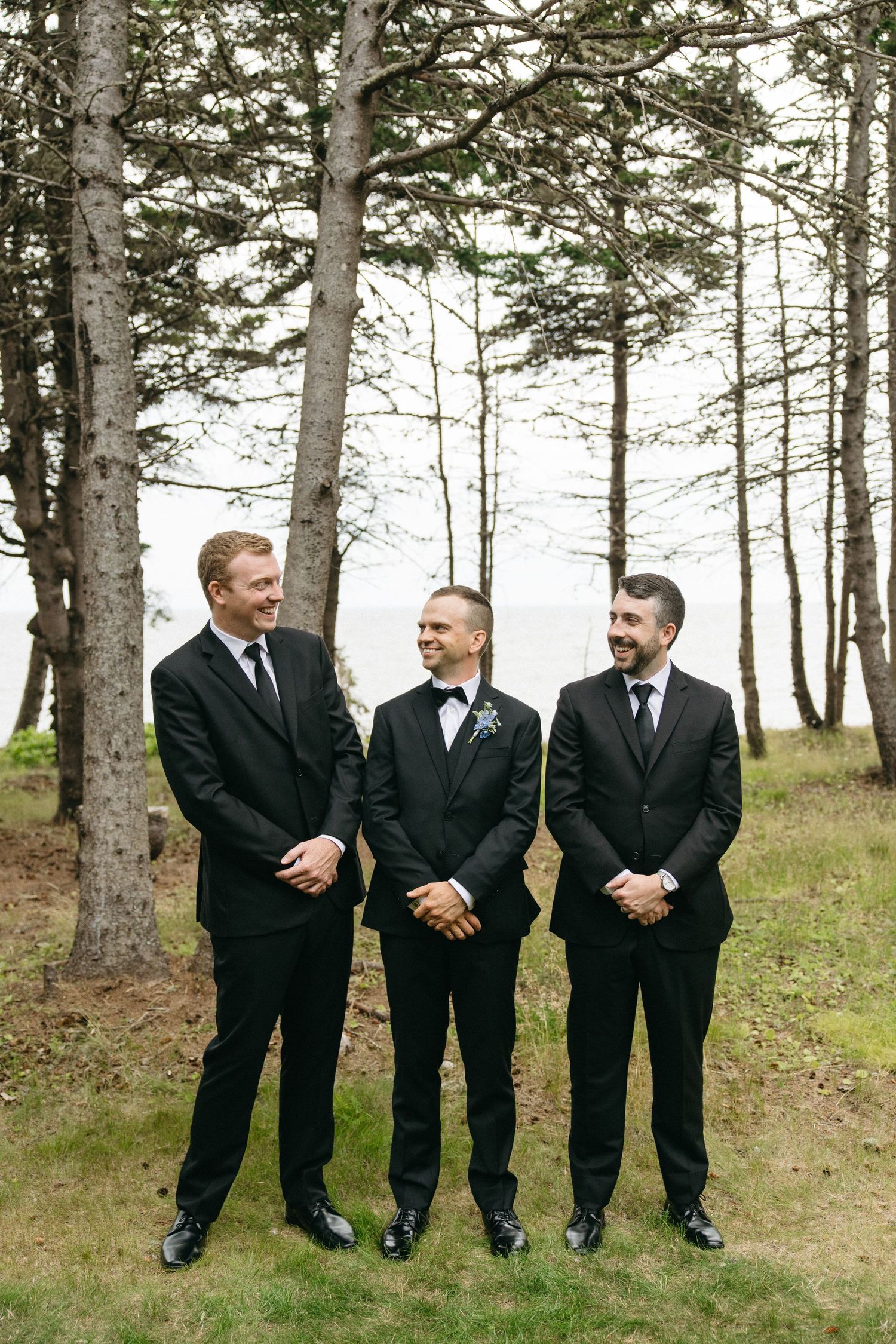 Three groomsmen in black tuxedos smiling outdoors among tall pine trees at a wedding.