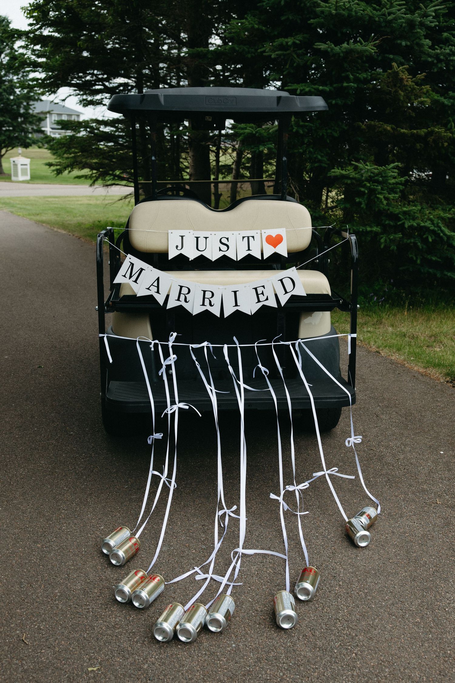 Golf cart decorated with Just Married banner and tin cans trailing white ribbons on a tree-lined path.