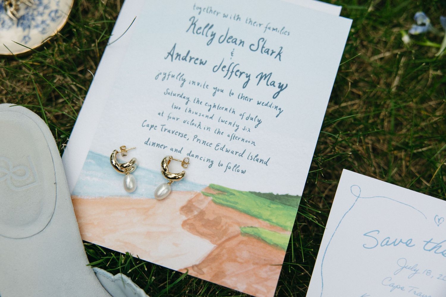 Elegant watercolor wedding invitation for Kelly and Andrew with coastal landscape design and pearl earrings on grass.