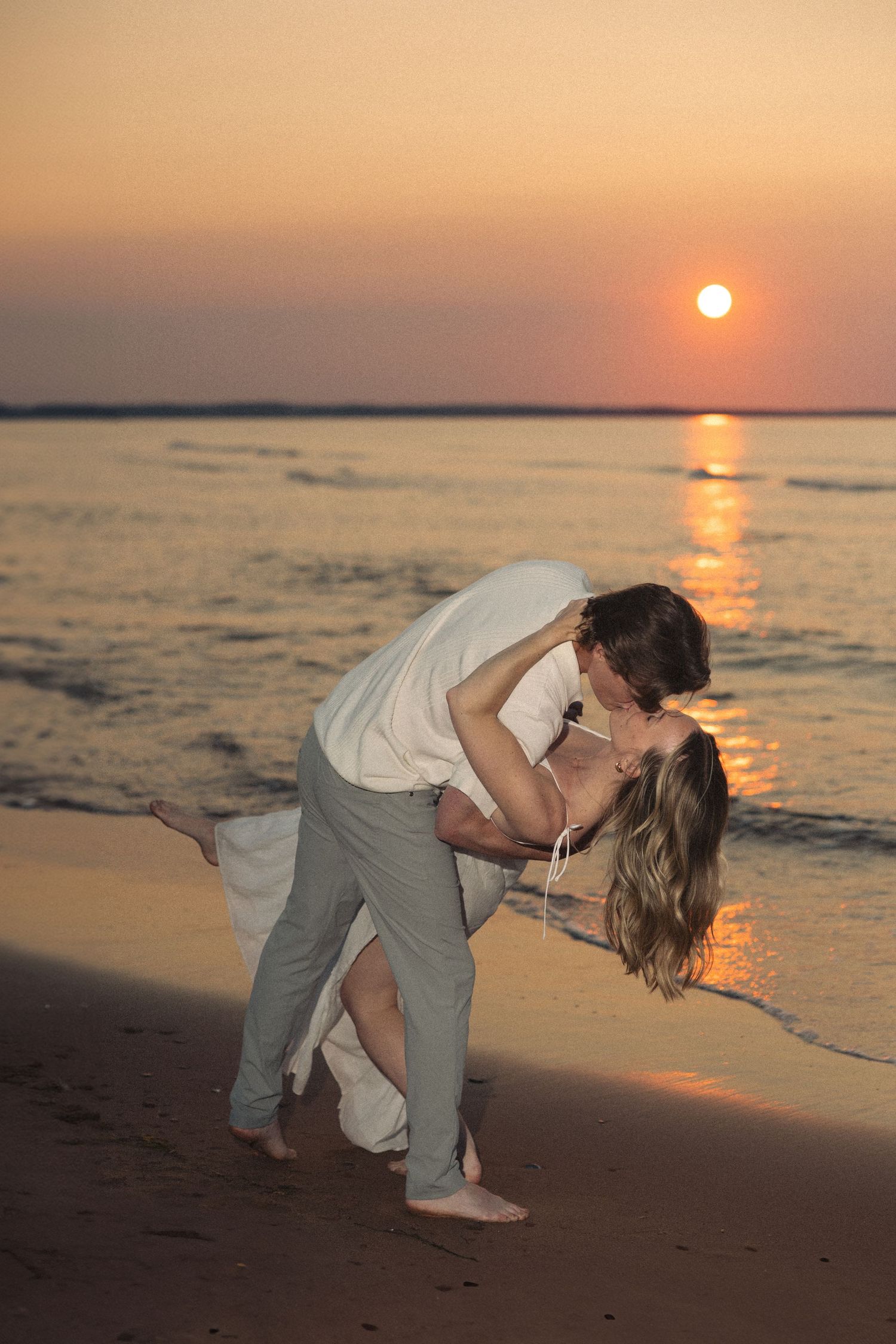 Couple sharing a romantic dip kiss on the beach at sunset with golden ocean backdrop.