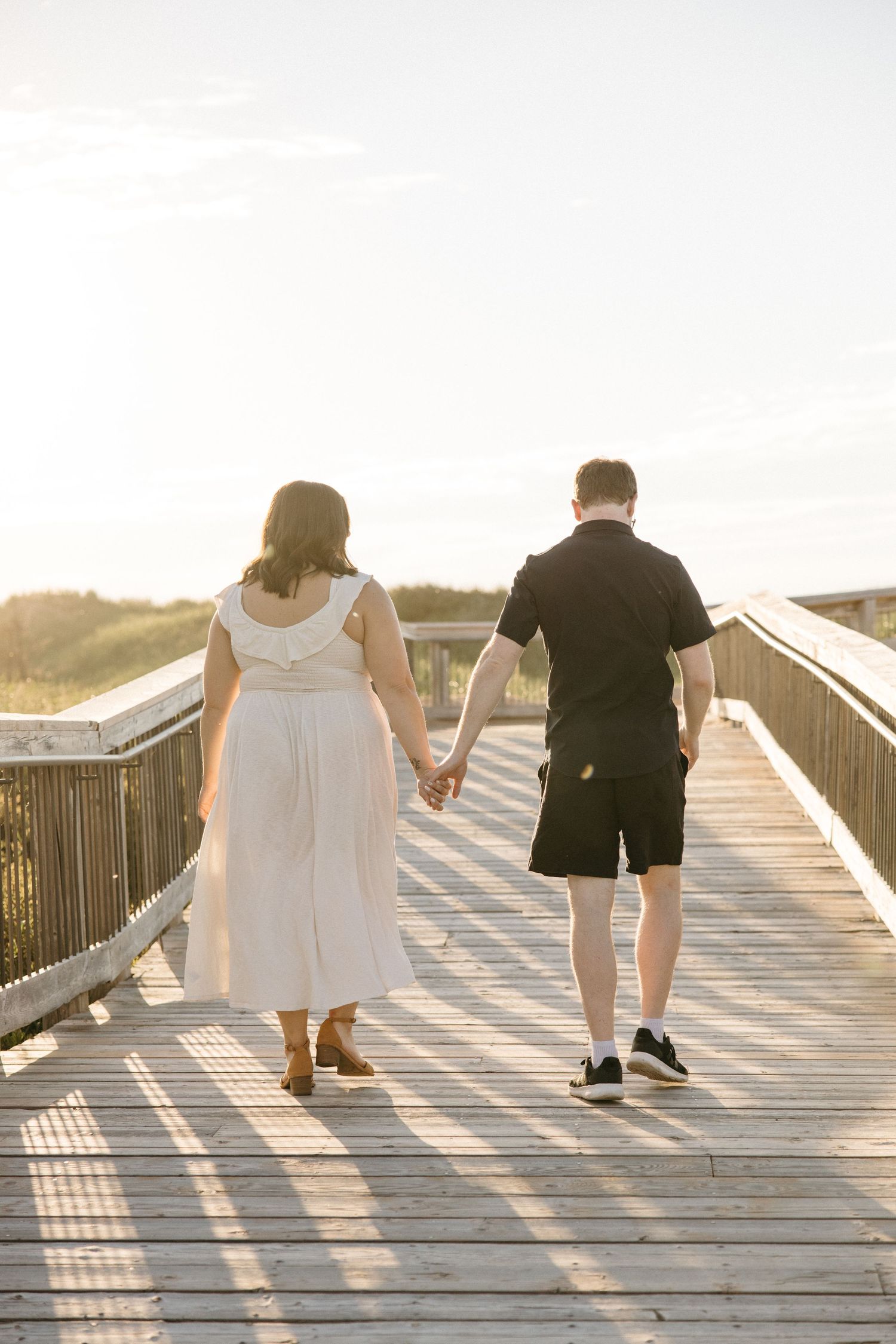 Couple holding hands walking away on wooden boardwalk toward bright sunset at beach.