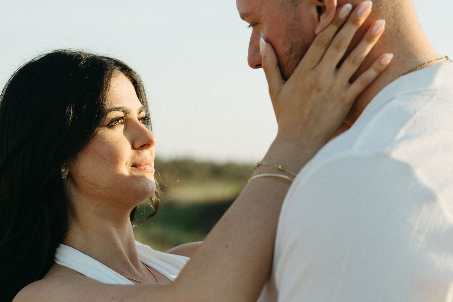 A woman tenderly holds a man's face while gazing up at him outdoors in warm sunlight, wearing white outfits.