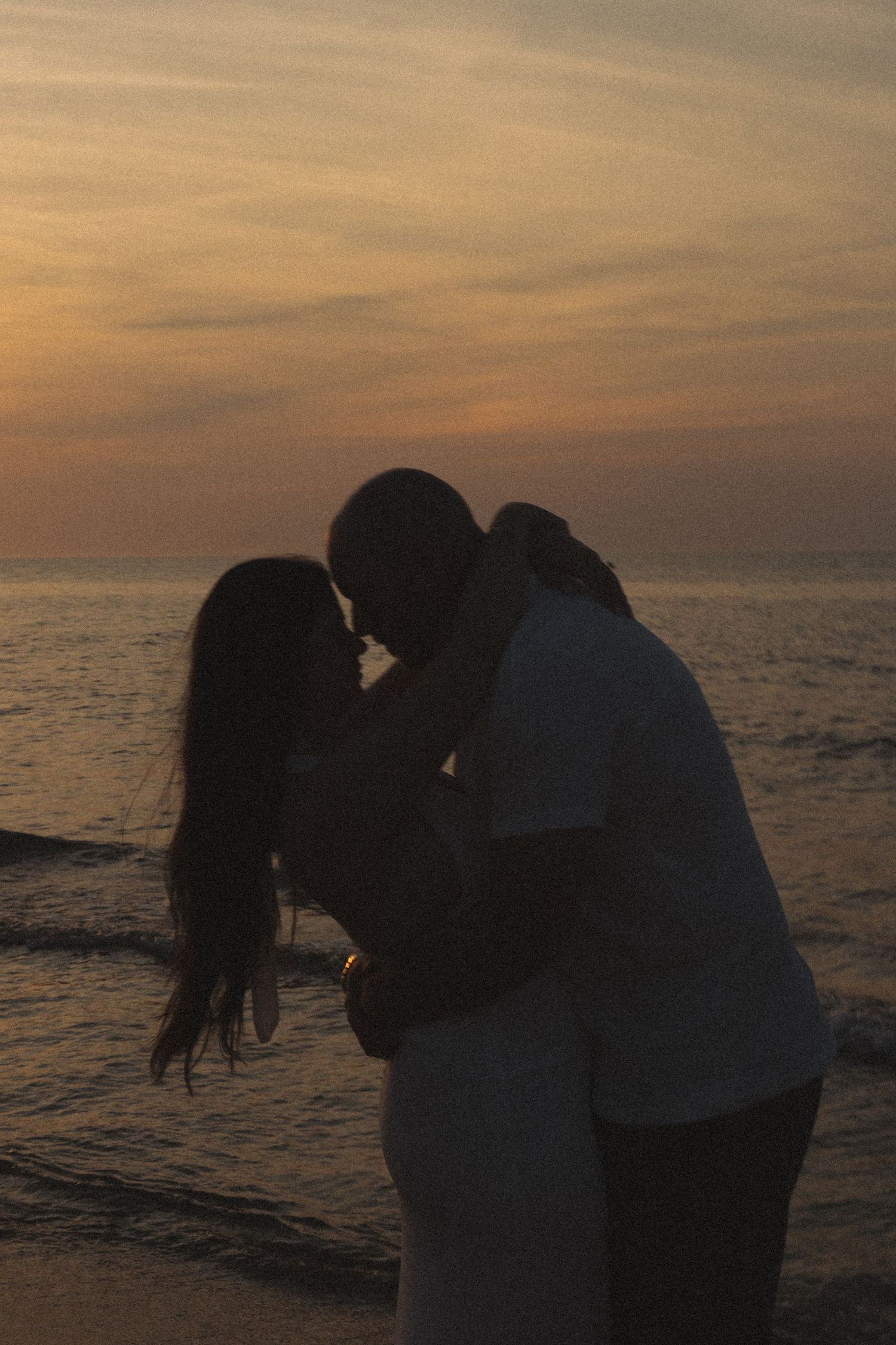Romantic couple silhouetted against vibrant orange sunset sky while kissing on a beach.