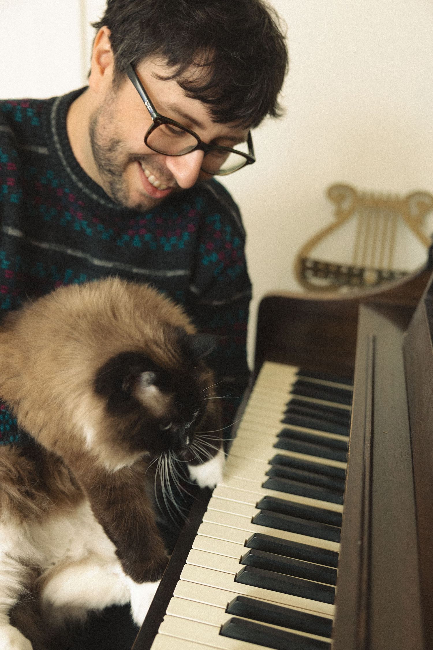Smiling man with glasses plays piano while a Siamese cat sits on the keys beside him.