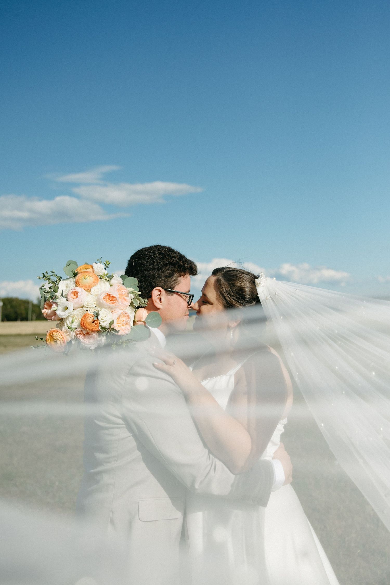 Bride and groom kissing under flowing veil against bright blue sky.