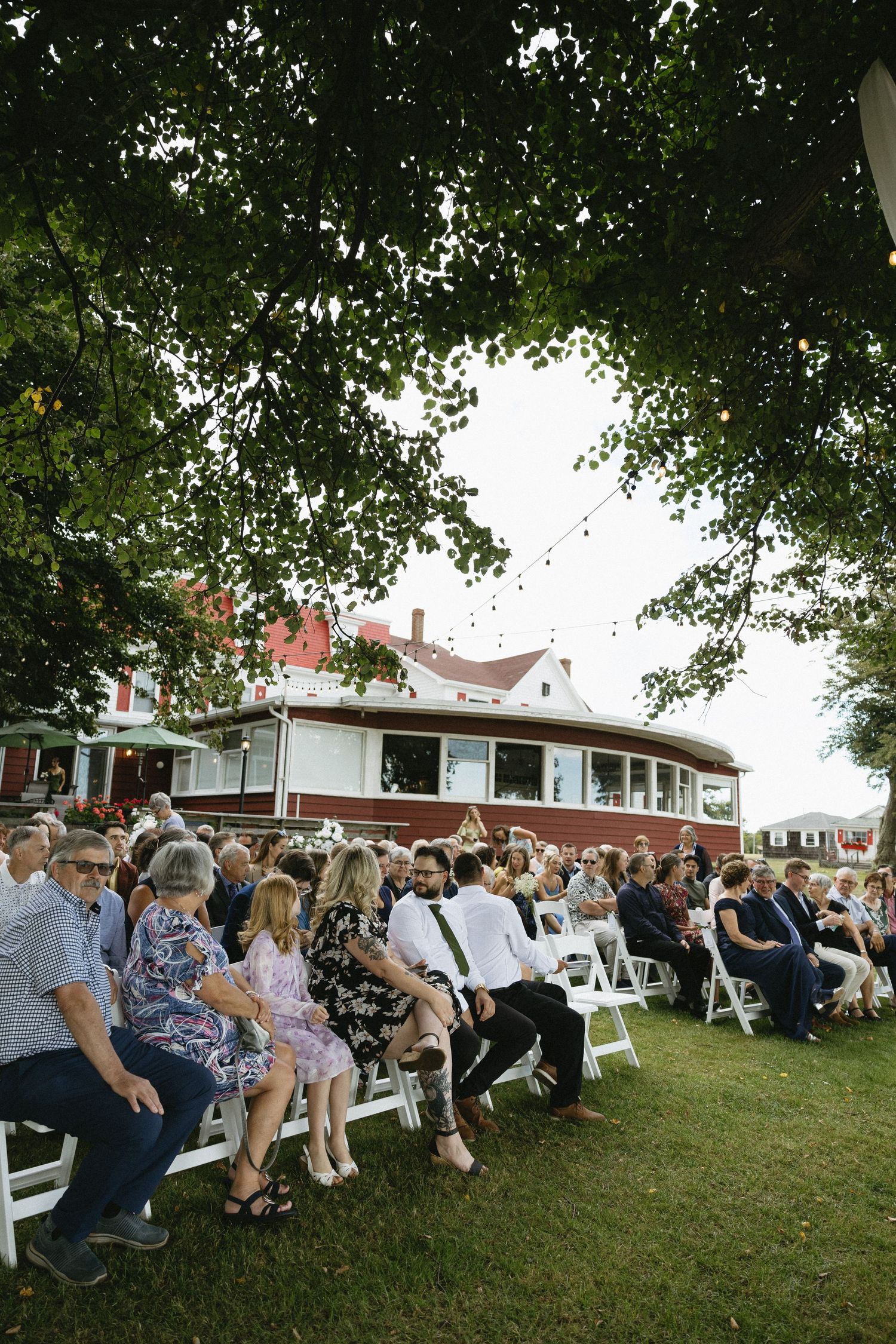 Wedding guests seated in white chairs at outdoor ceremony with red venue building visible in background.