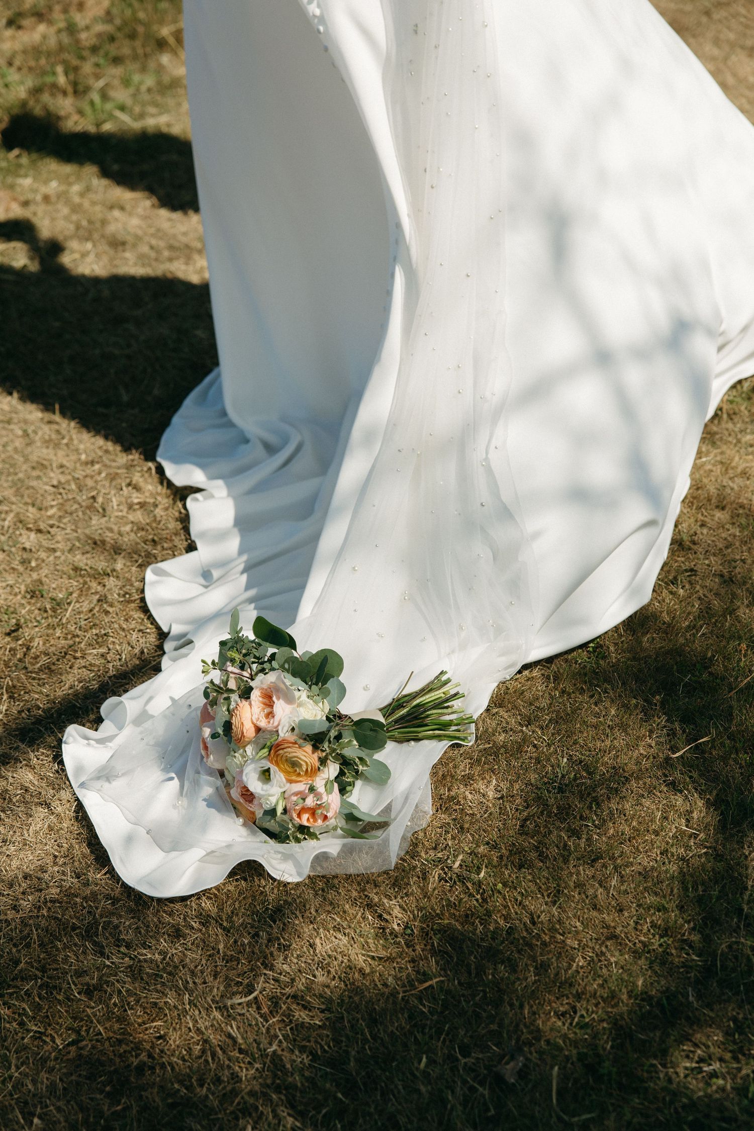 Bride in flowing white wedding gown holding a peach and white floral bouquet on dry grass.