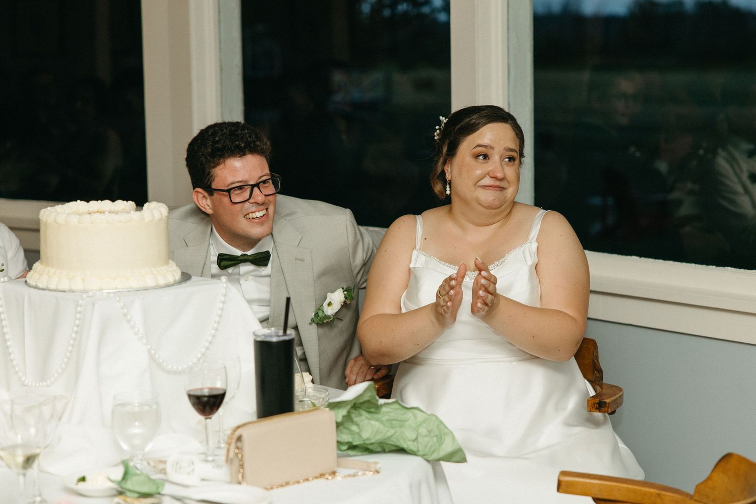 Bride and groom seated at wedding reception table, smiling and clapping near a white tiered cake.