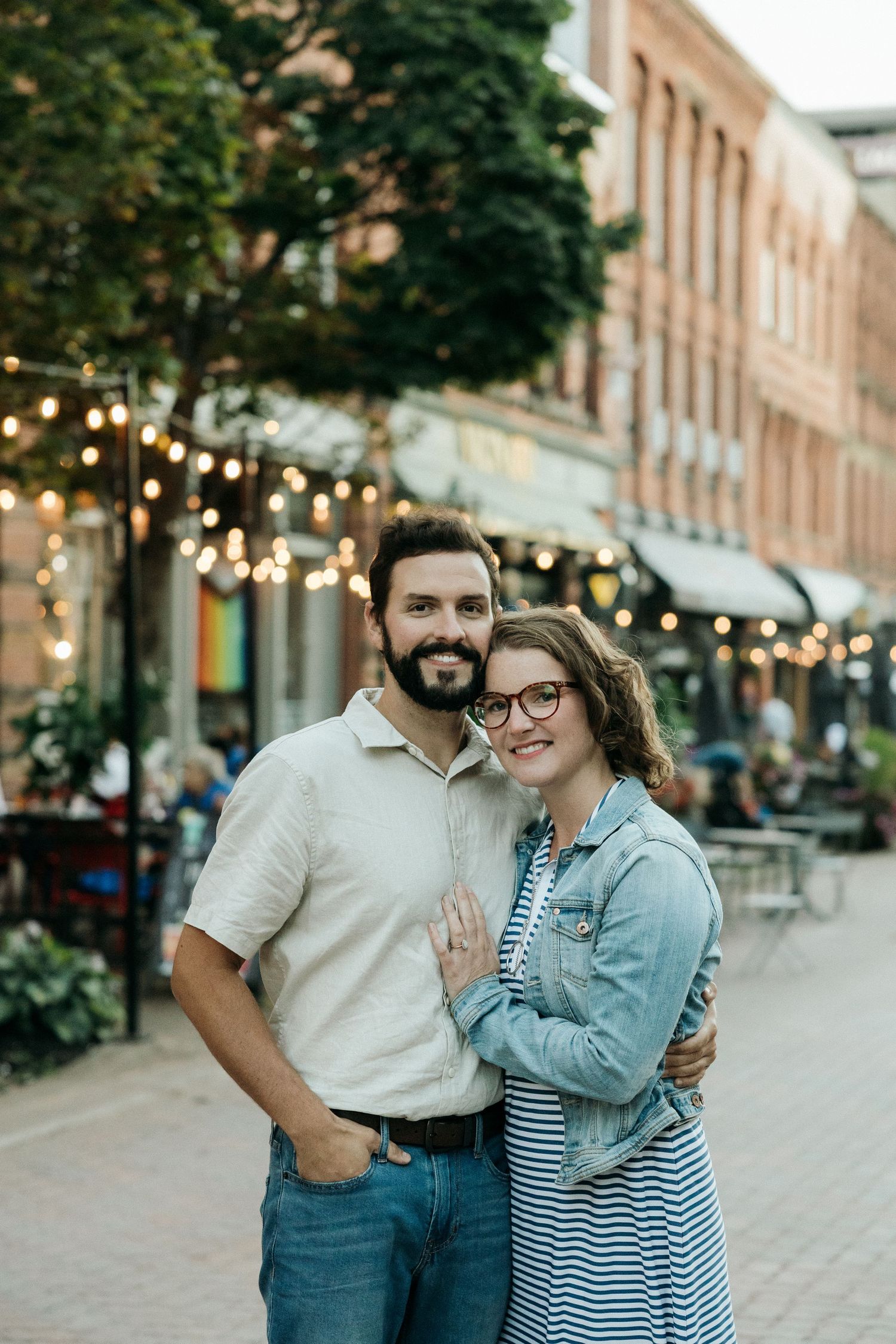 A couple shares a romantic moment on a charming city street with string lights and brick buildings in the background.