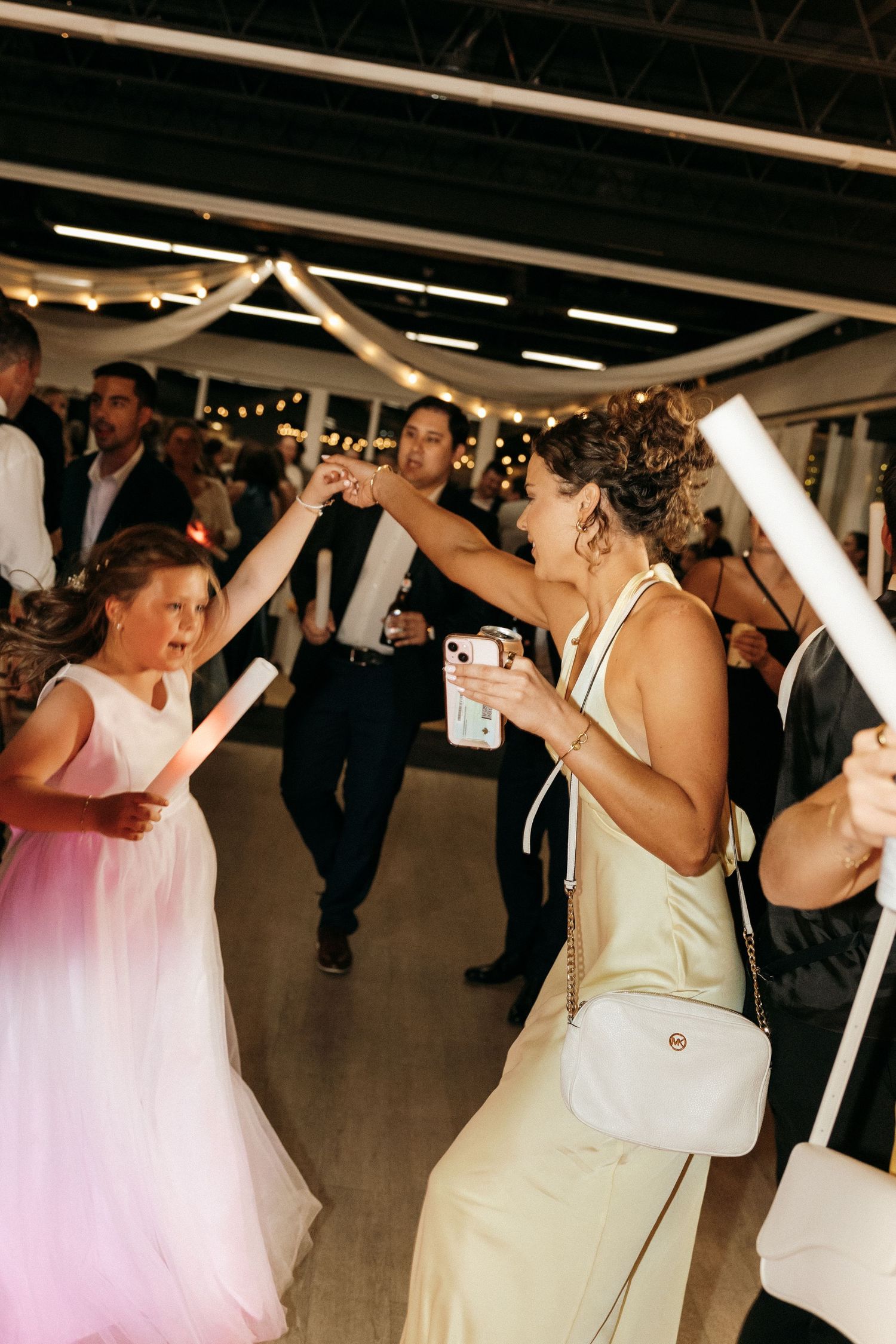 Wedding guests dance with raised arms under string lights at an indoor reception venue.