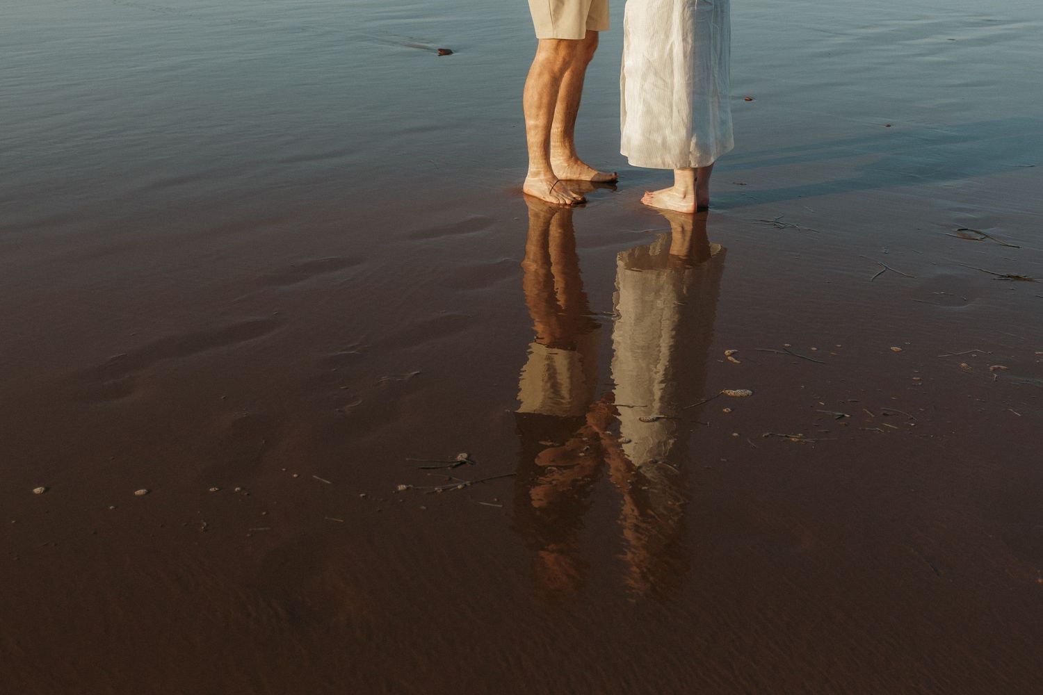 Two people stand ankle-deep in calm ocean waters at sunset with their reflections visible in the wet sand.