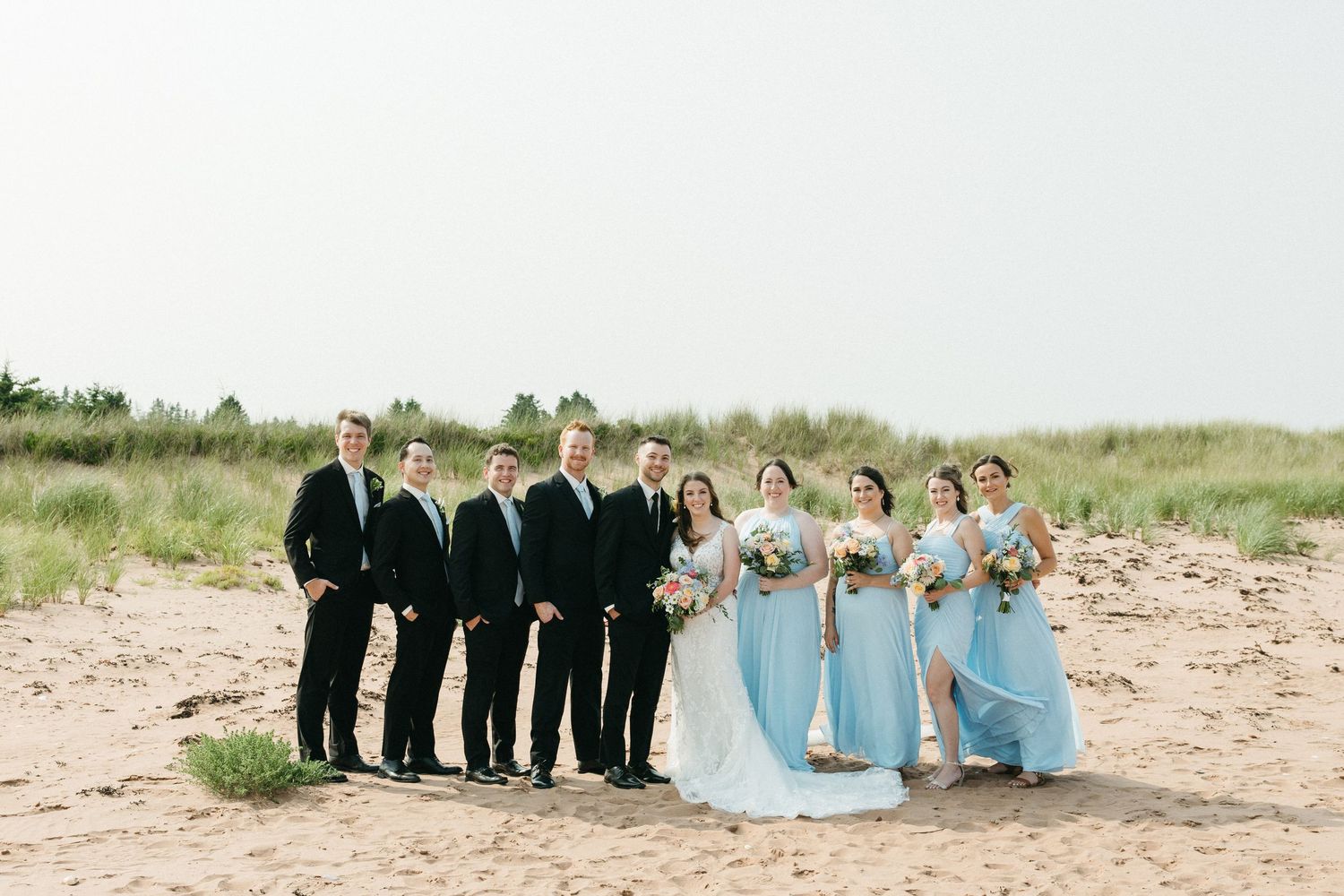 Wedding party in black suits and light blue dresses poses together on a sandy beach.