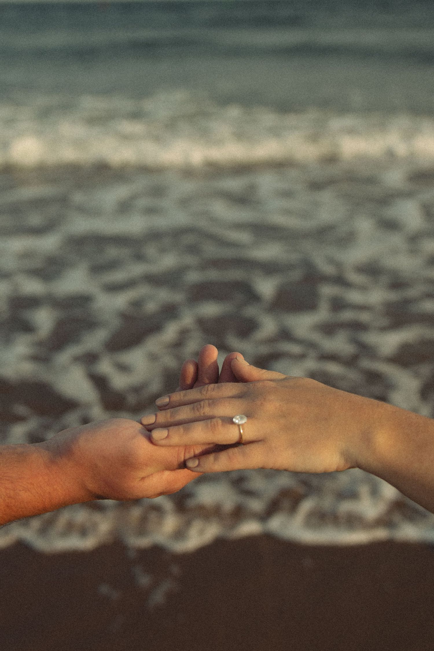 Close-up of hands reaching towards each other with an engagement ring against an ocean wave background.