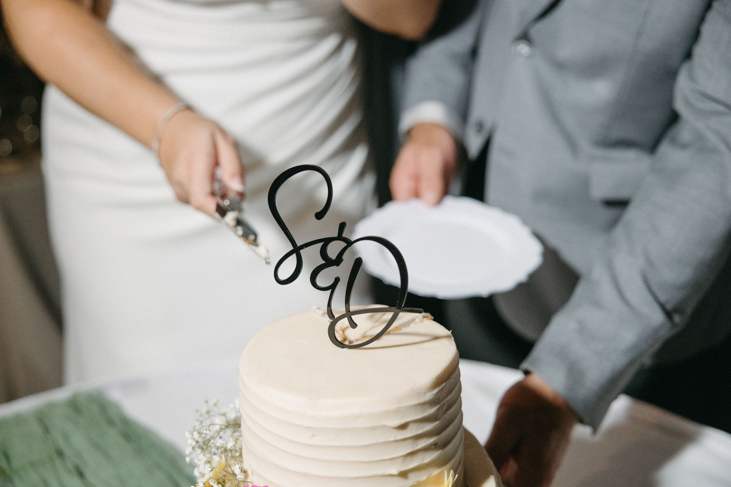 Close-up of hands cutting into a white wedding cake decorated with fresh flowers.