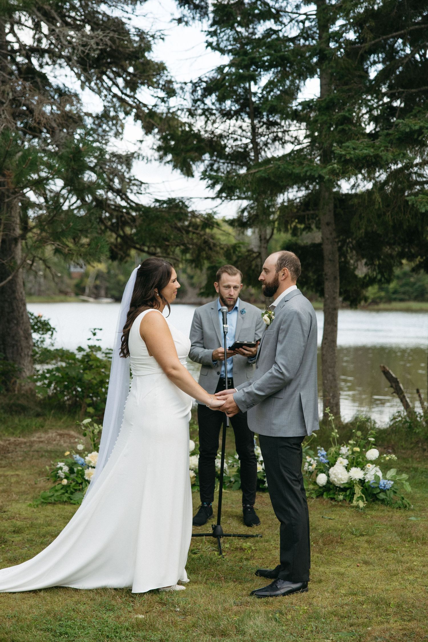 Outdoor wedding ceremony taking place by a lake with evergreen trees in background.