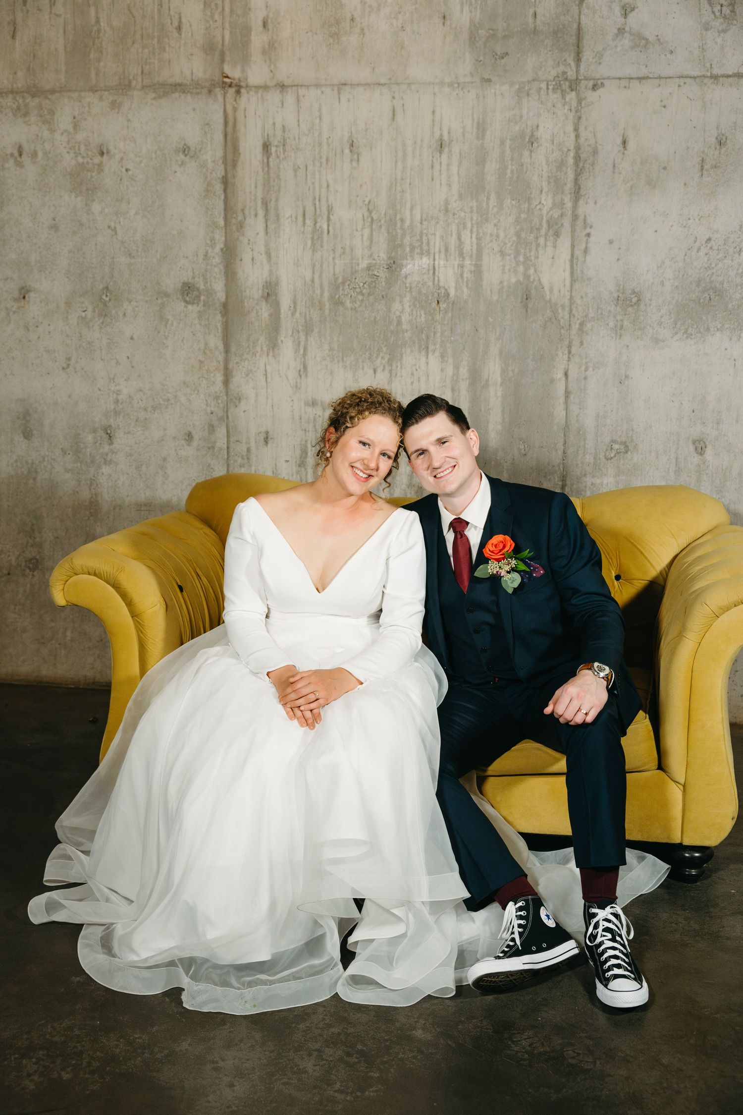 Couple in wedding attire relaxes on yellow couch, showing off casual sneakers.