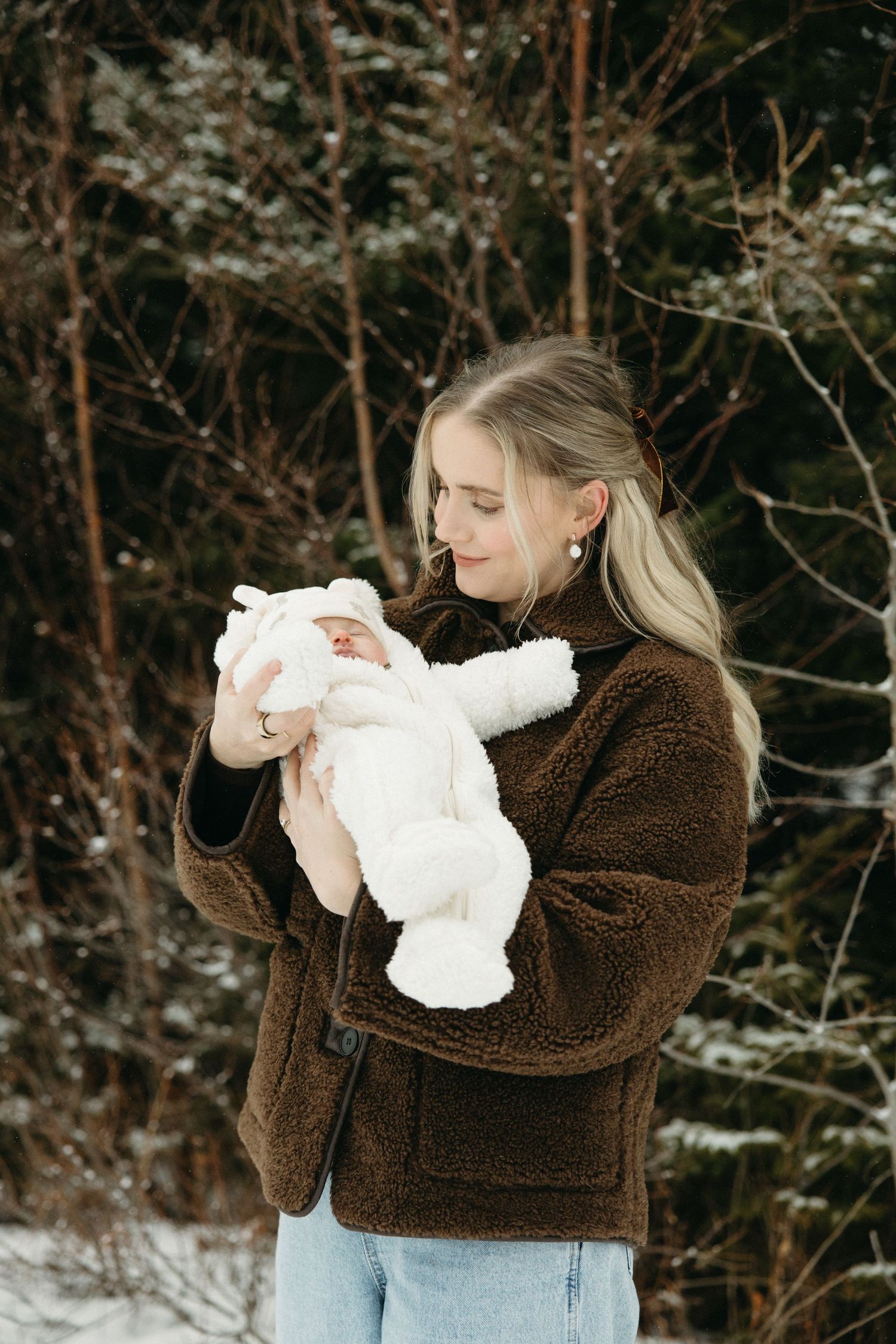 Mother in brown jacket holding baby dressed in white snowsuit during winter photoshoot.