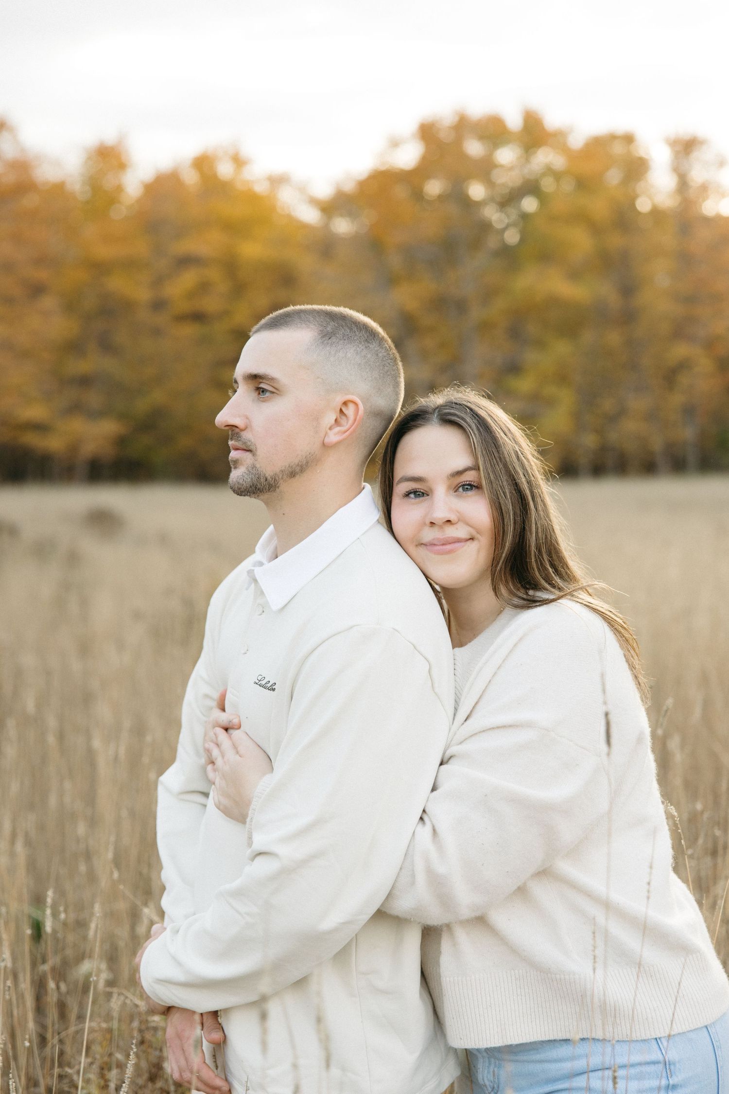 Romantic couple in light colored outfits pose together in rustic field during autumn sunset.