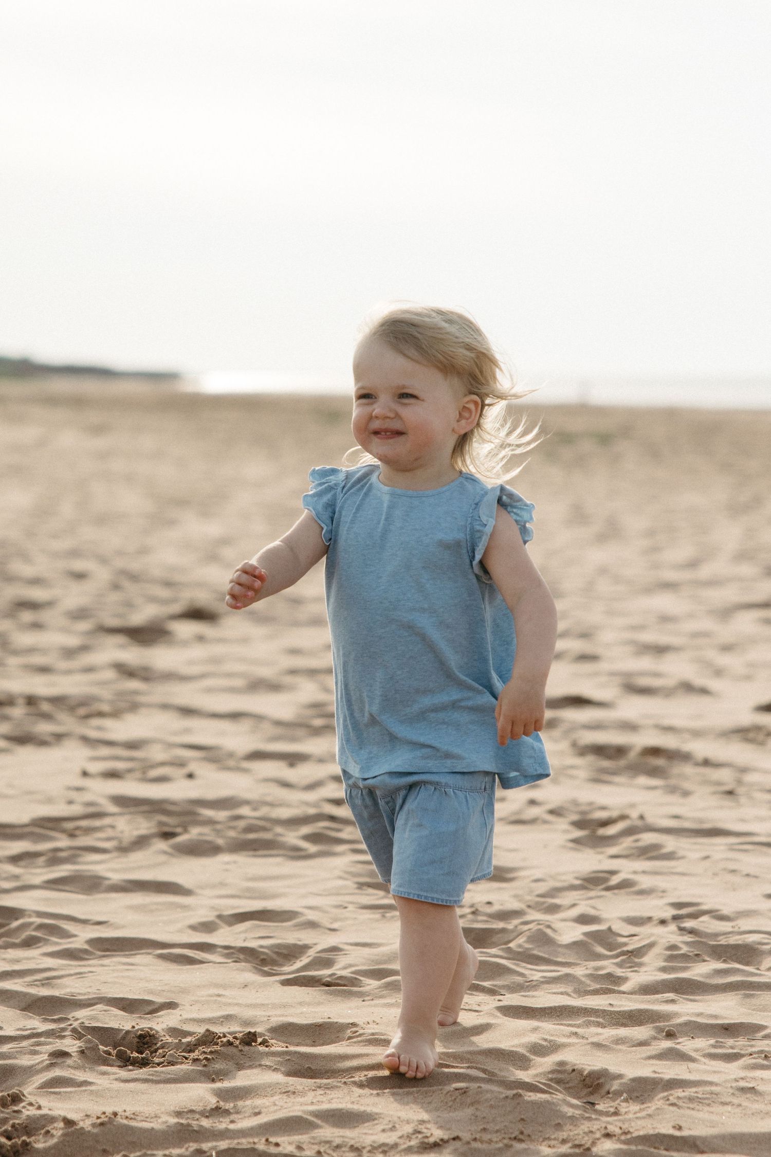 A happy blonde toddler girl in a blue outfit runs barefoot along a sandy beach.