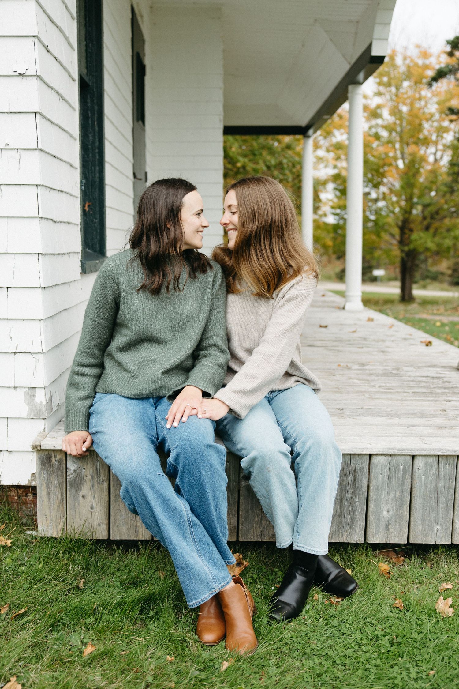 Two women in jeans and sweaters sit close on a white porch edge, smiling at each other, with autumn trees and green grass behind them.