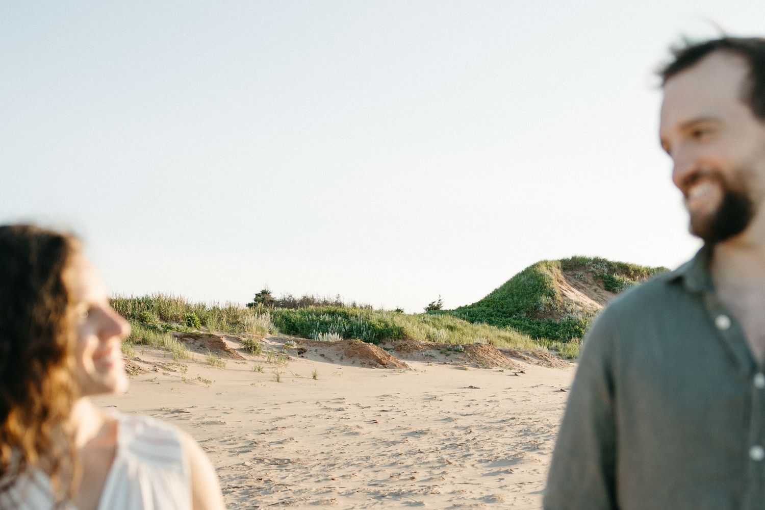 Couple smiling on a sandy beach with green dunes and clear sky in the background.