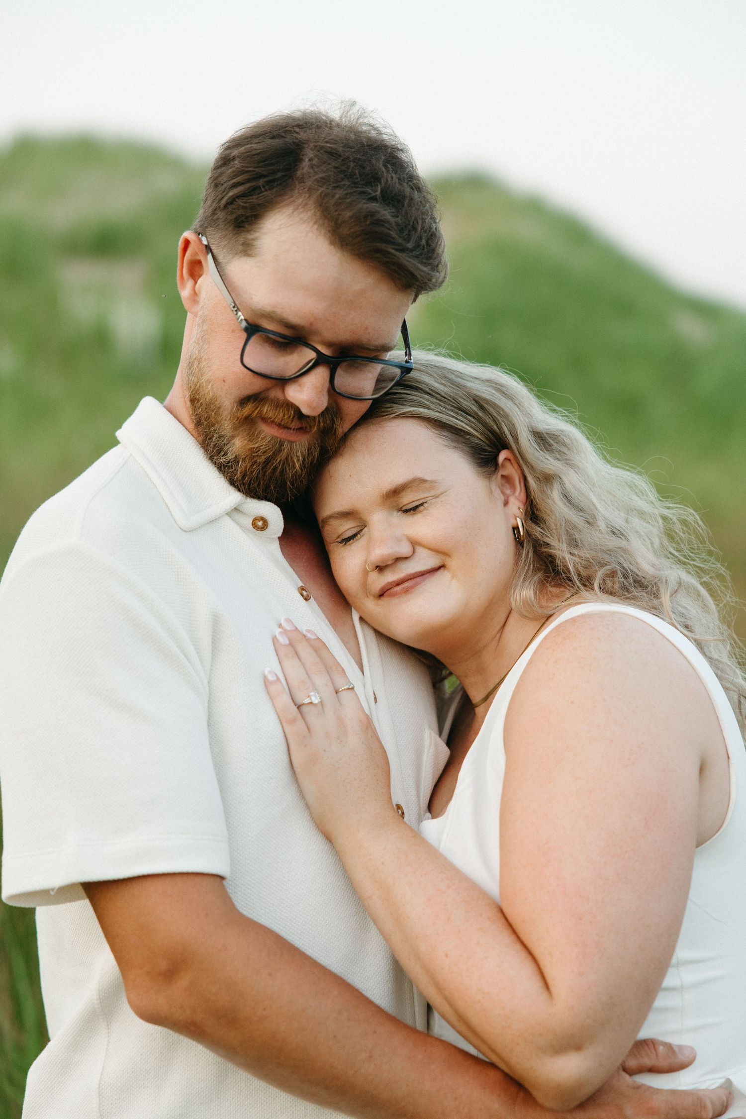 Couple embracing tenderly in green meadow, woman showing engagement ring while leaning on mans chest.