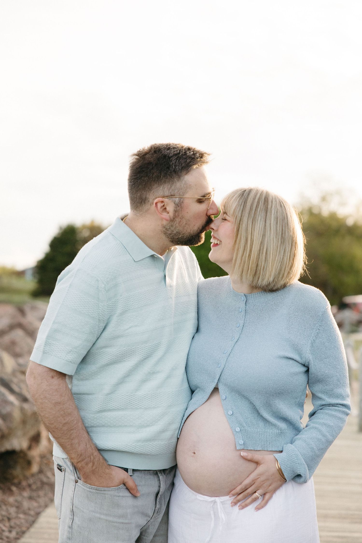 Expecting couple sharing a tender kiss on a boardwalk, man holding pregnant woman's belly in matching blue outfits.