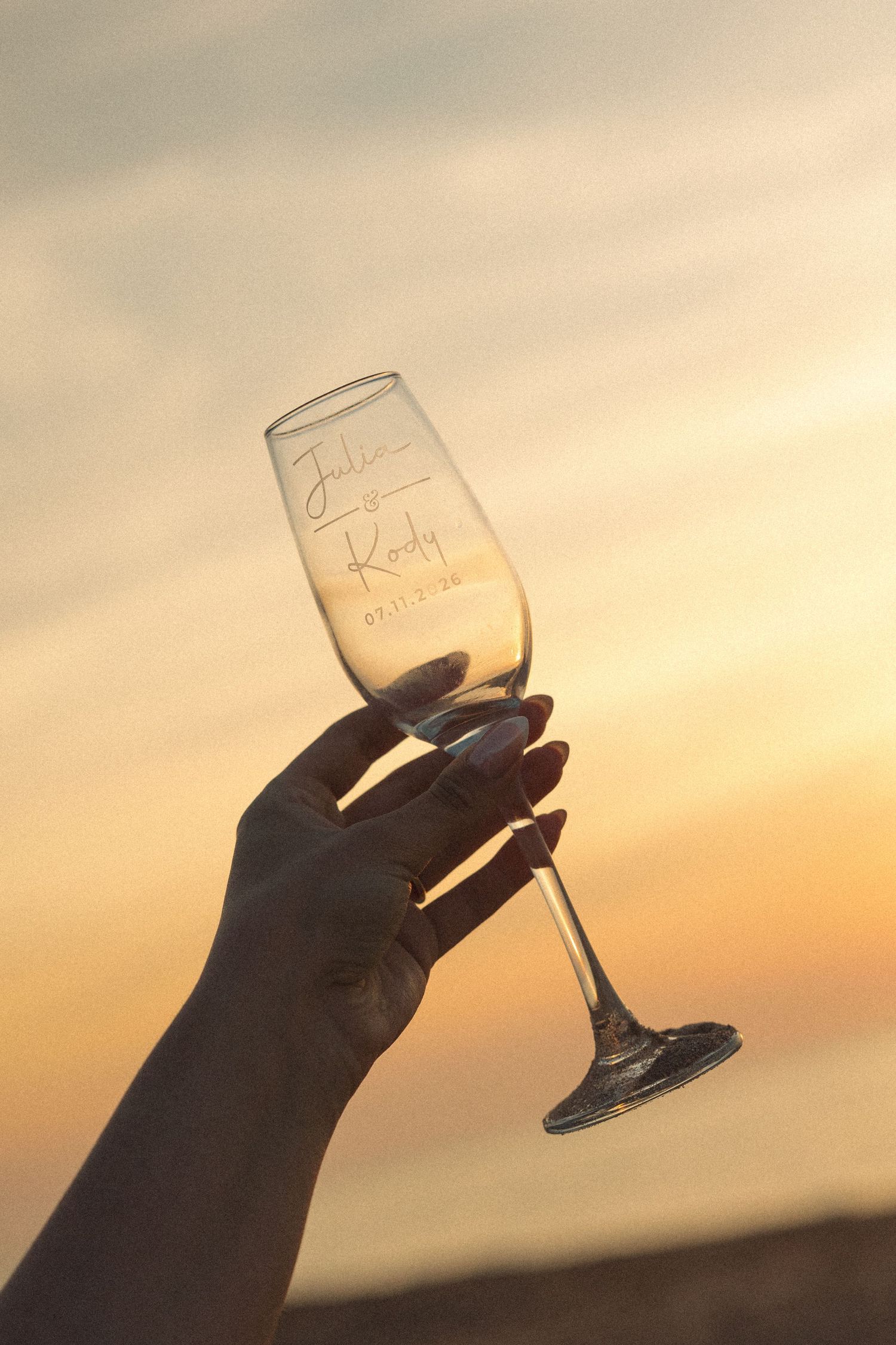 Hand holding personalized engraved champagne flute with names against a warm sunset sky.