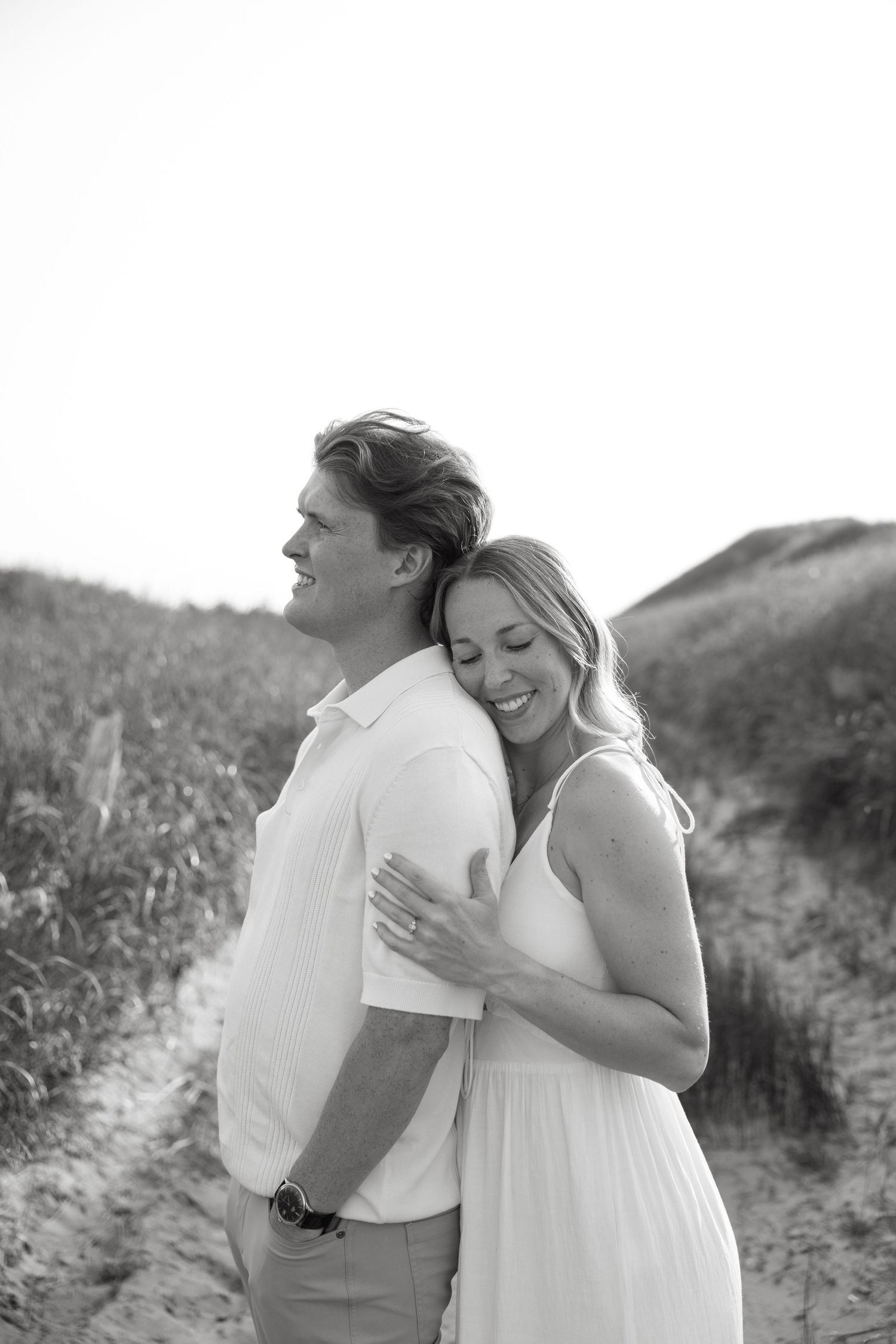 Black and white photo of a couple embracing on a sandy beach path, woman in white dress smiling with eyes closed.