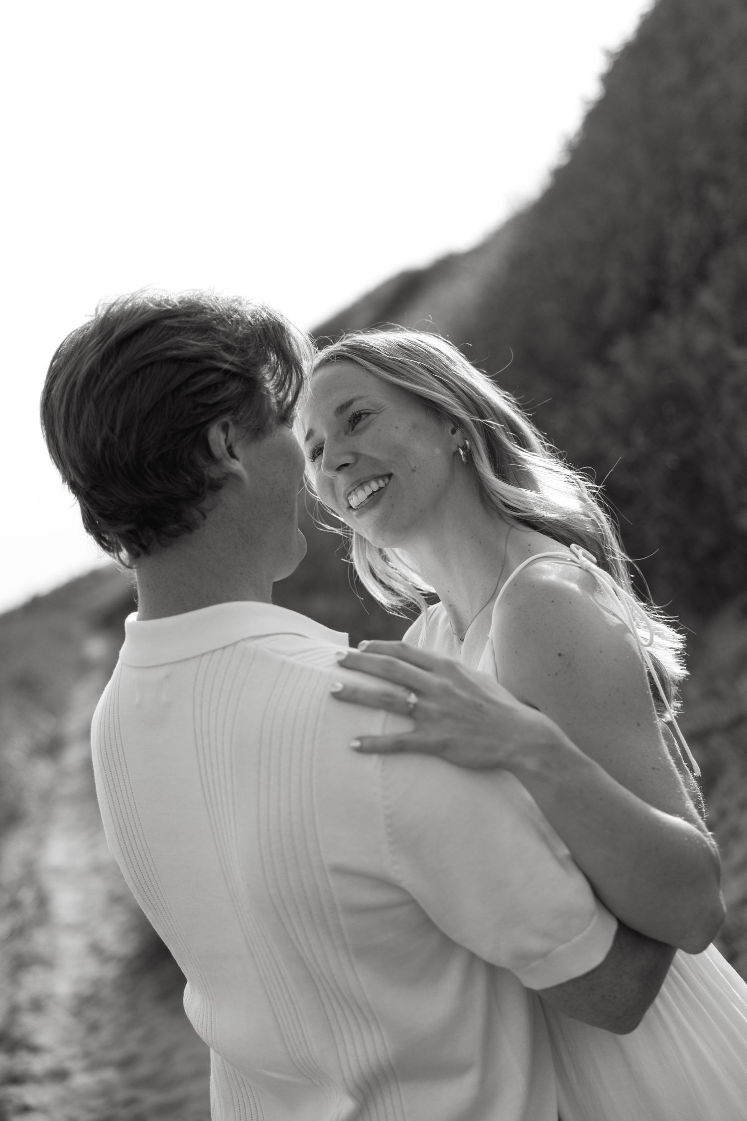 Black and white photo of laughing woman embracing man outdoors near grassy dunes in romantic moment.