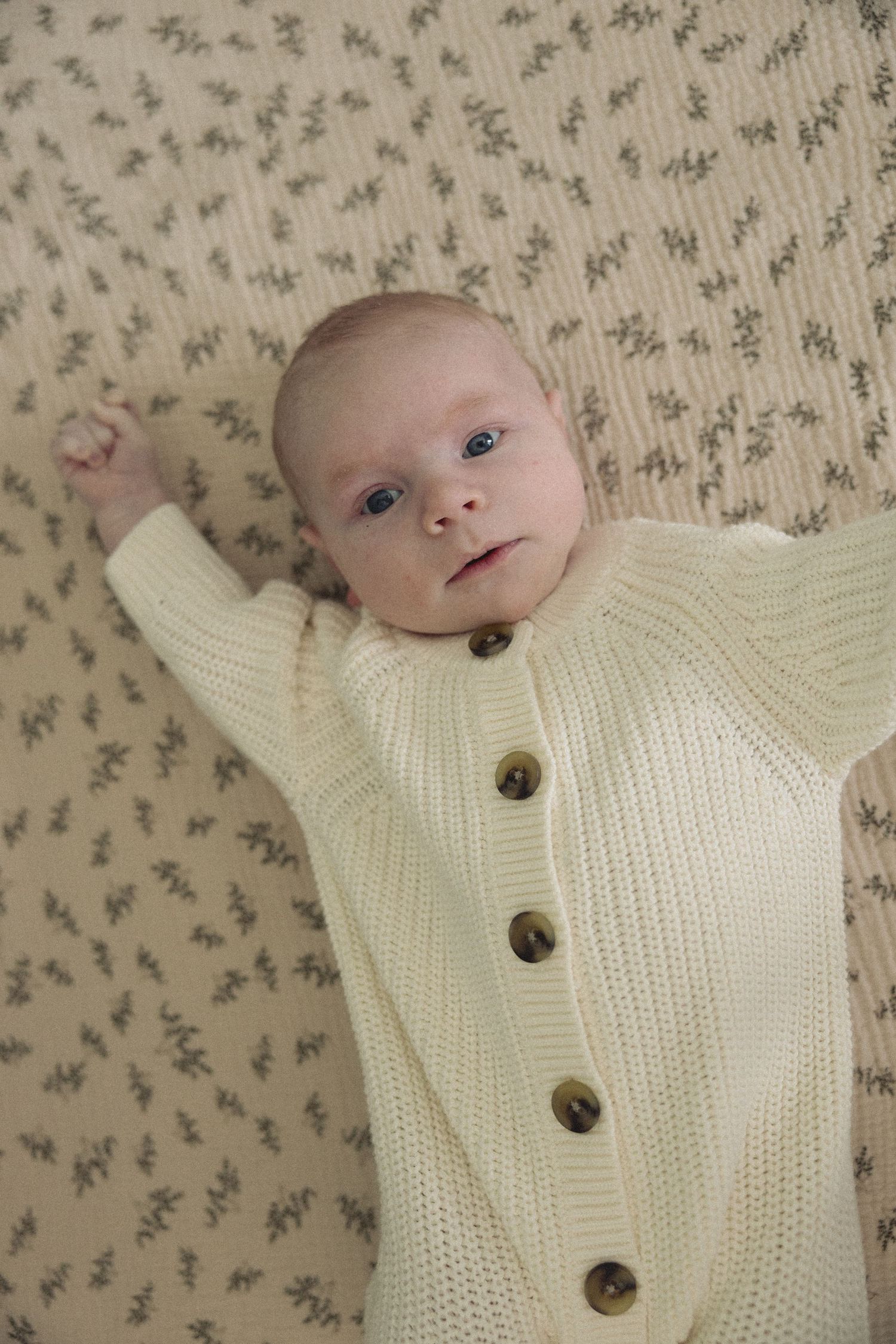 Baby wearing cream knit cardigan with buttons lies on floral pattern fabric.